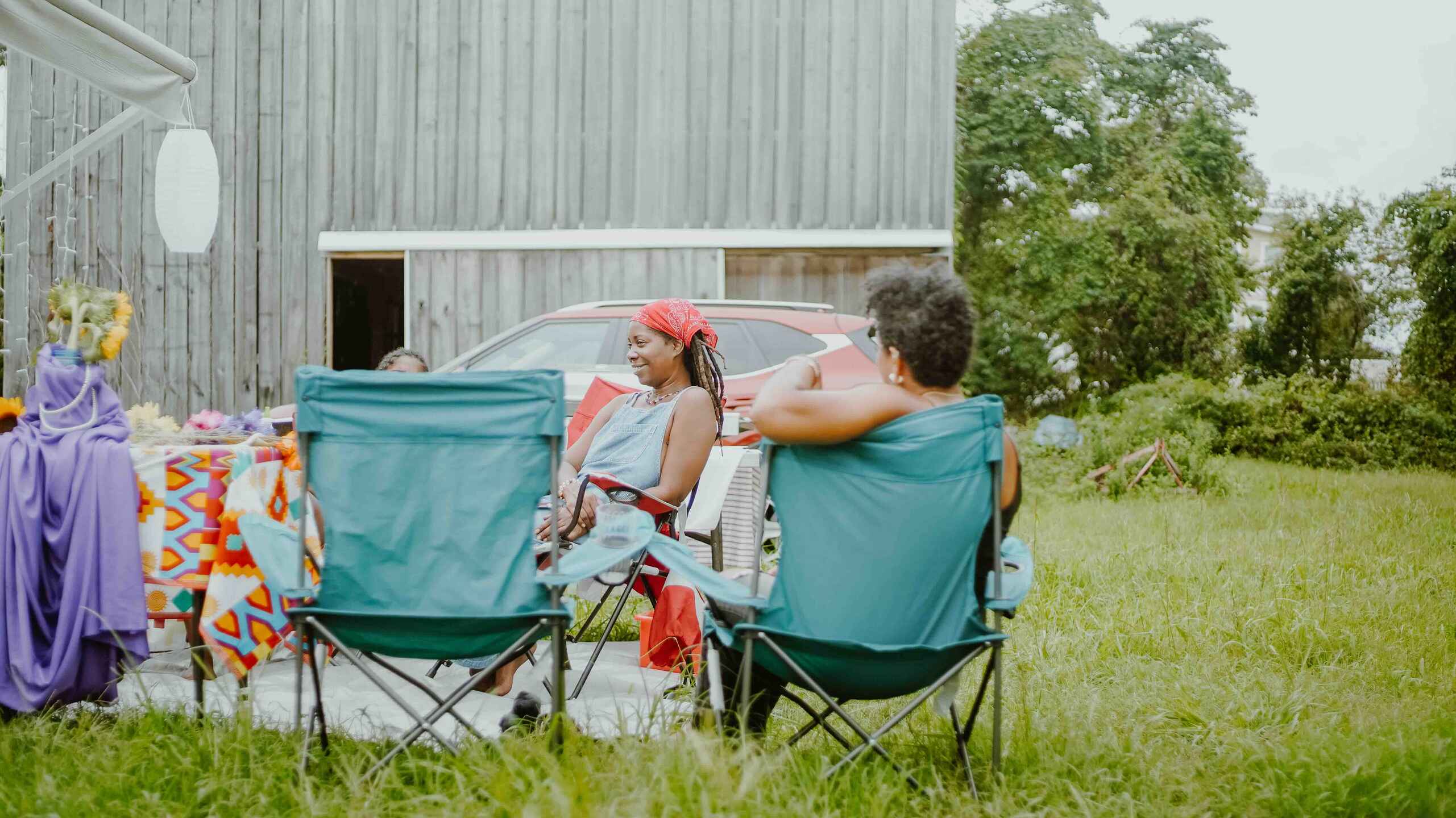 Two people sit in camping chairs outside a farm building, enjoying a casual moment together