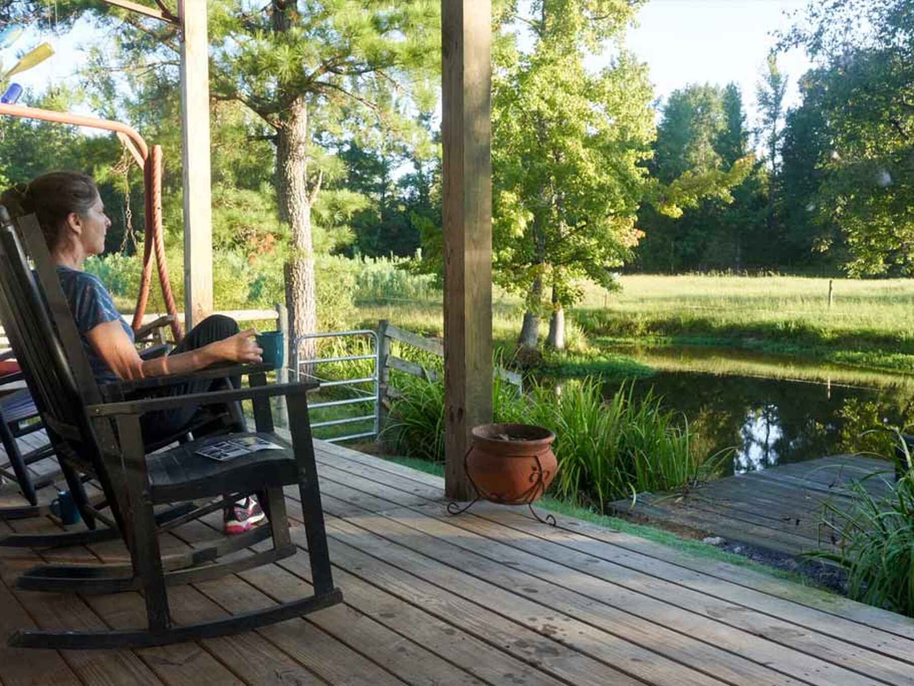 Person relaxing in a rocking chair on a wooden deck overlooking a pond surrounded by trees and farmland