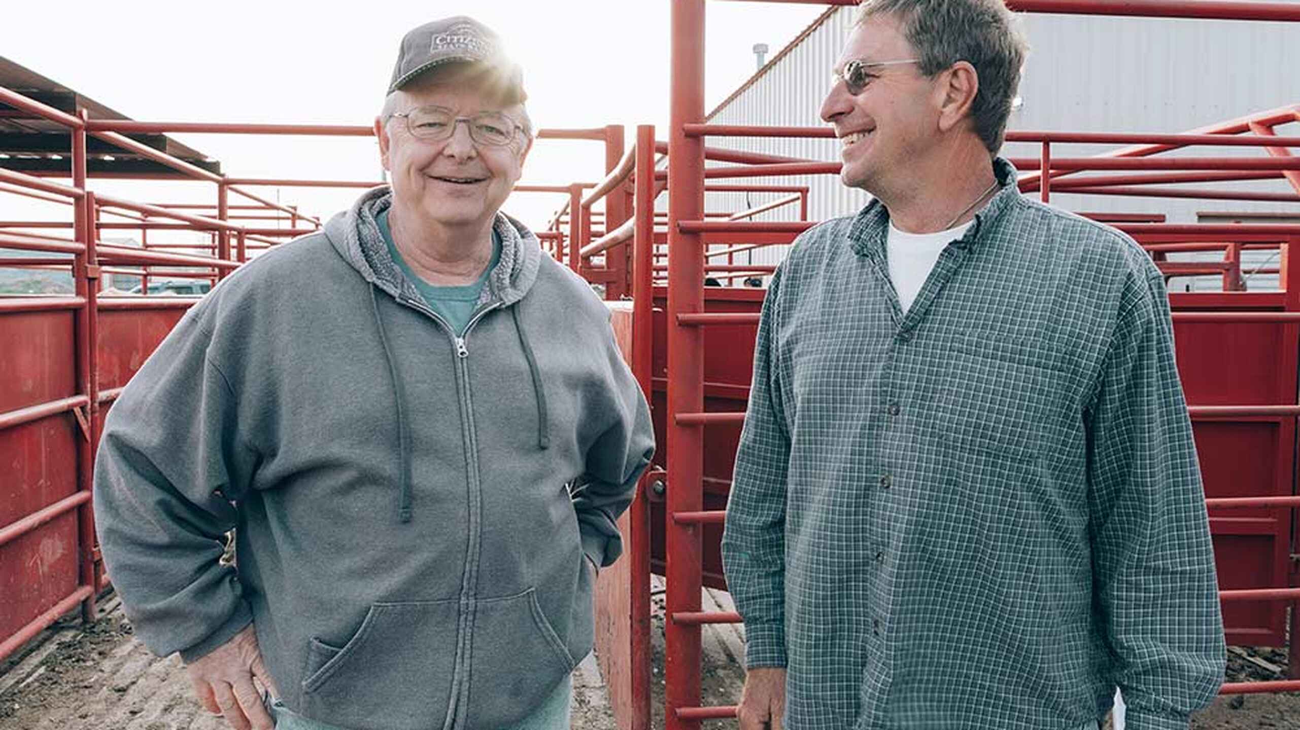Two farmers stand together in a livestock pen, smiling and conversing