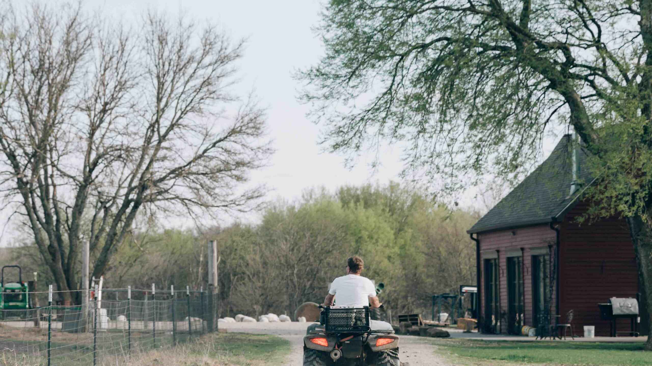 Farmer operating an ATV on a rural dirt road with farm buildings and bare trees in the background