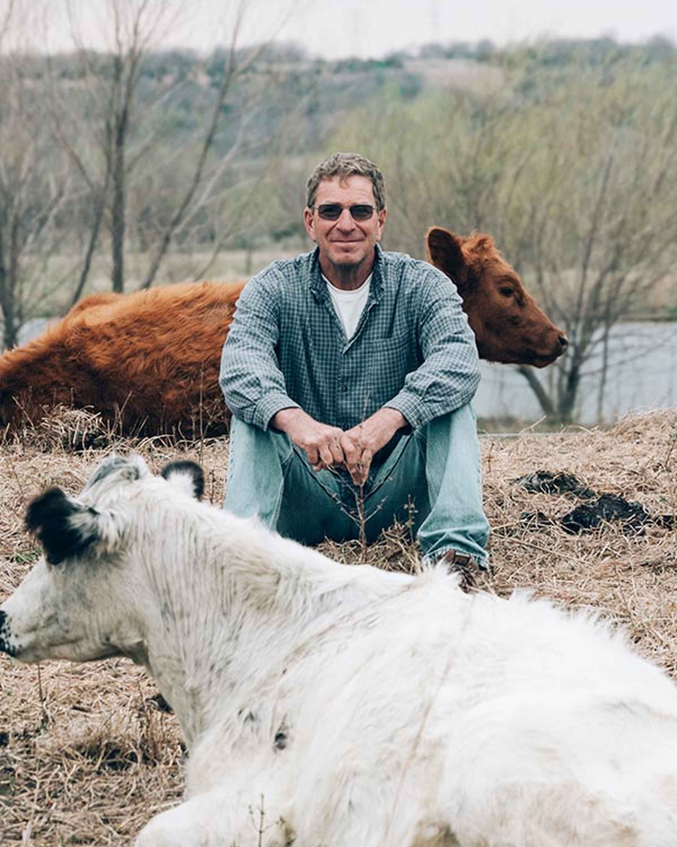Gail Fuller crouches among goats by a river with bare trees in the background
