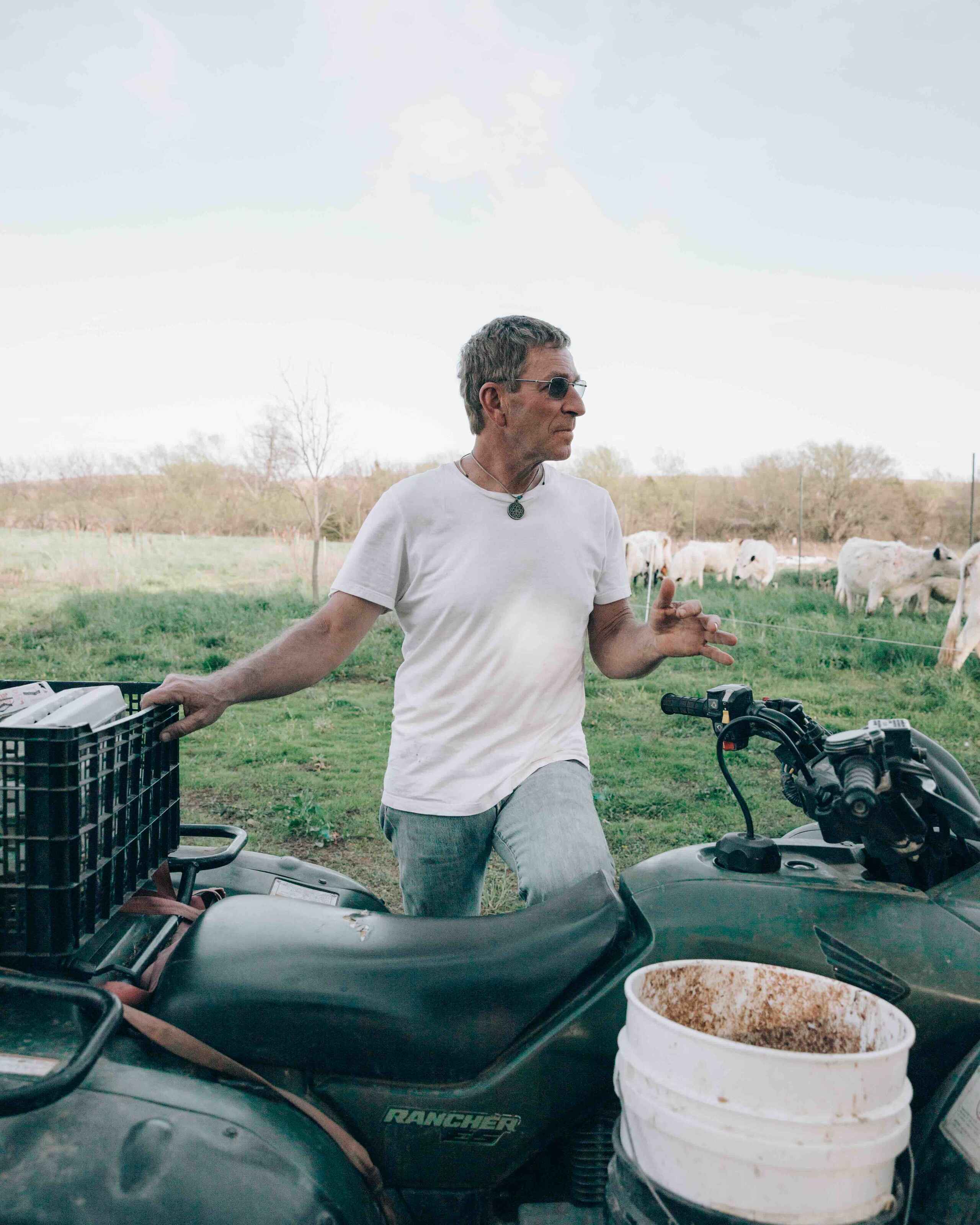 Gail Fuller stands on an ATV in a pasture while gesturing toward grazing cattle in the background