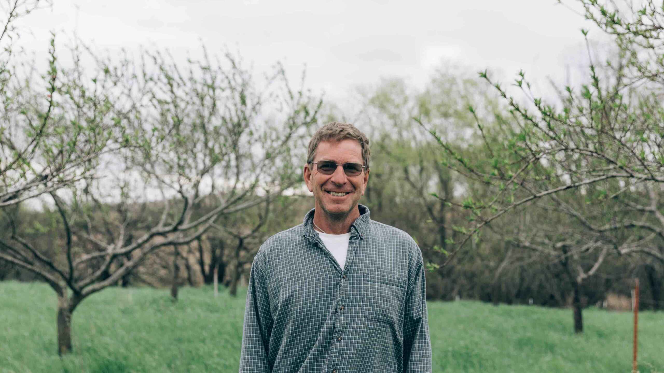 Gail Fuller standing in an orchard with young trees budding in spring