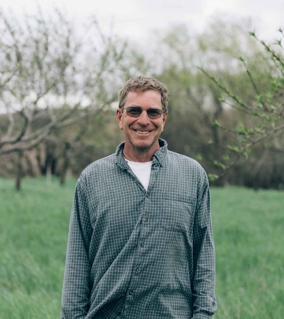 Gail Fuller stands smiling in an orchard wearing sunglasses and a gray plaid shirt