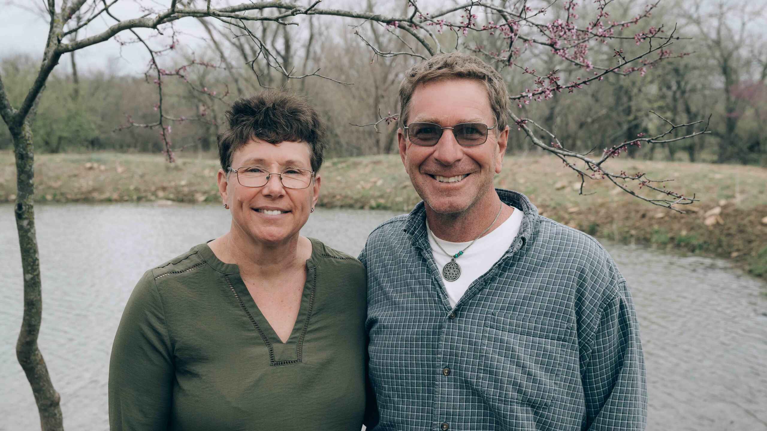 Gail Fuller and a male colleague stand together by a river with blooming trees in the background