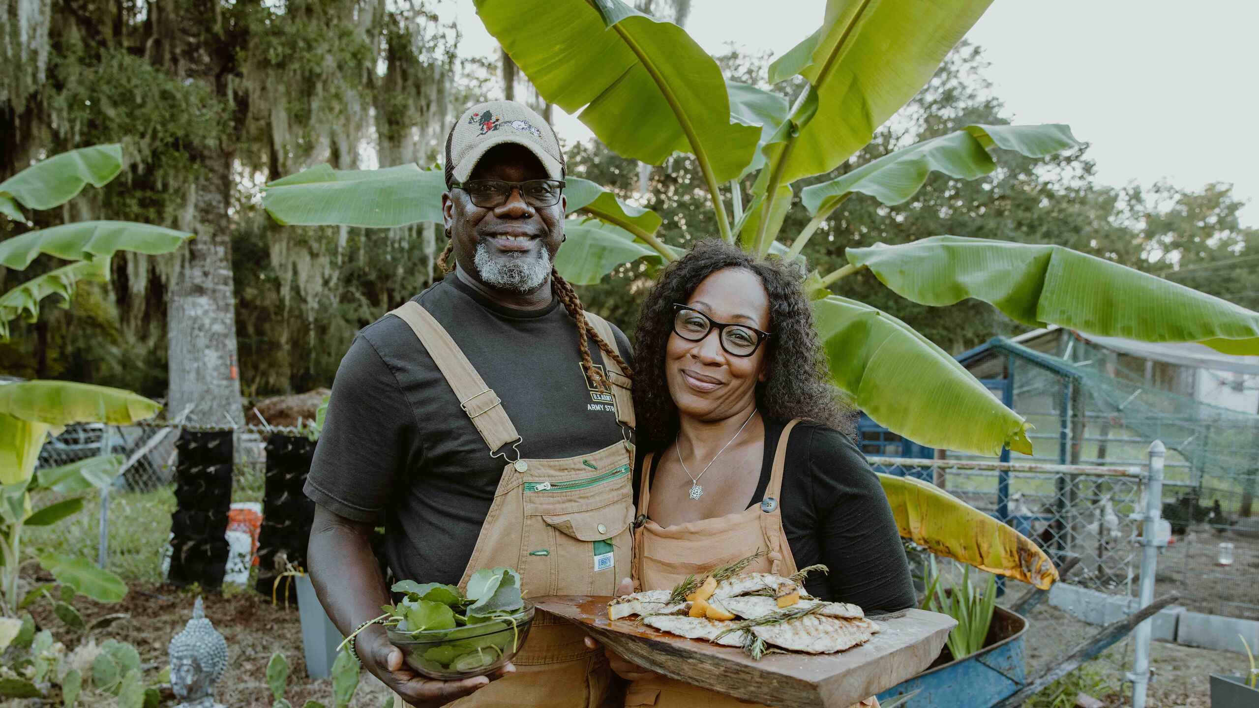 Two farmers holding fresh greens and fish in a garden with banana plants