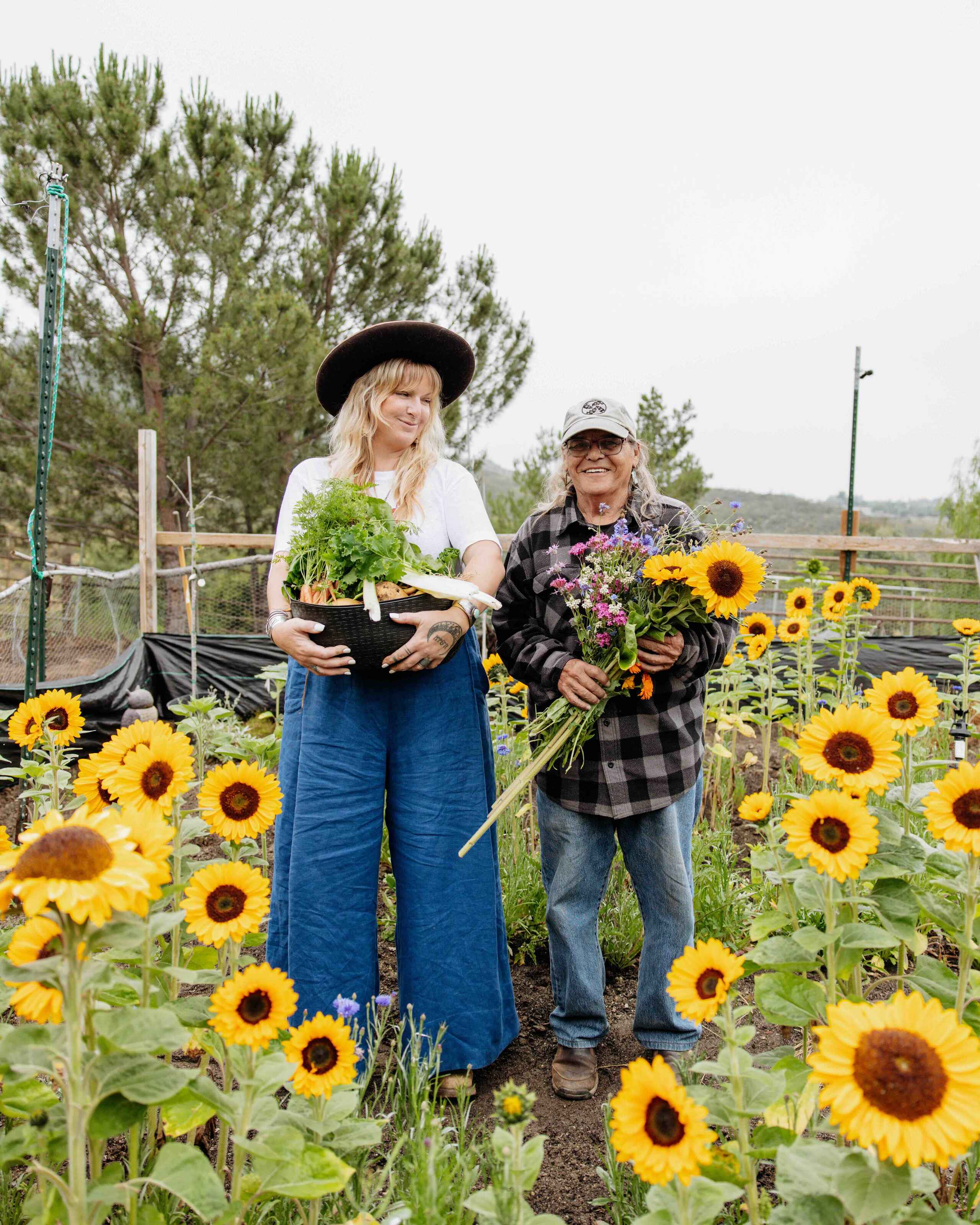 Two farmers holding fresh greens and sunflowers in a field of blooming sunflowers