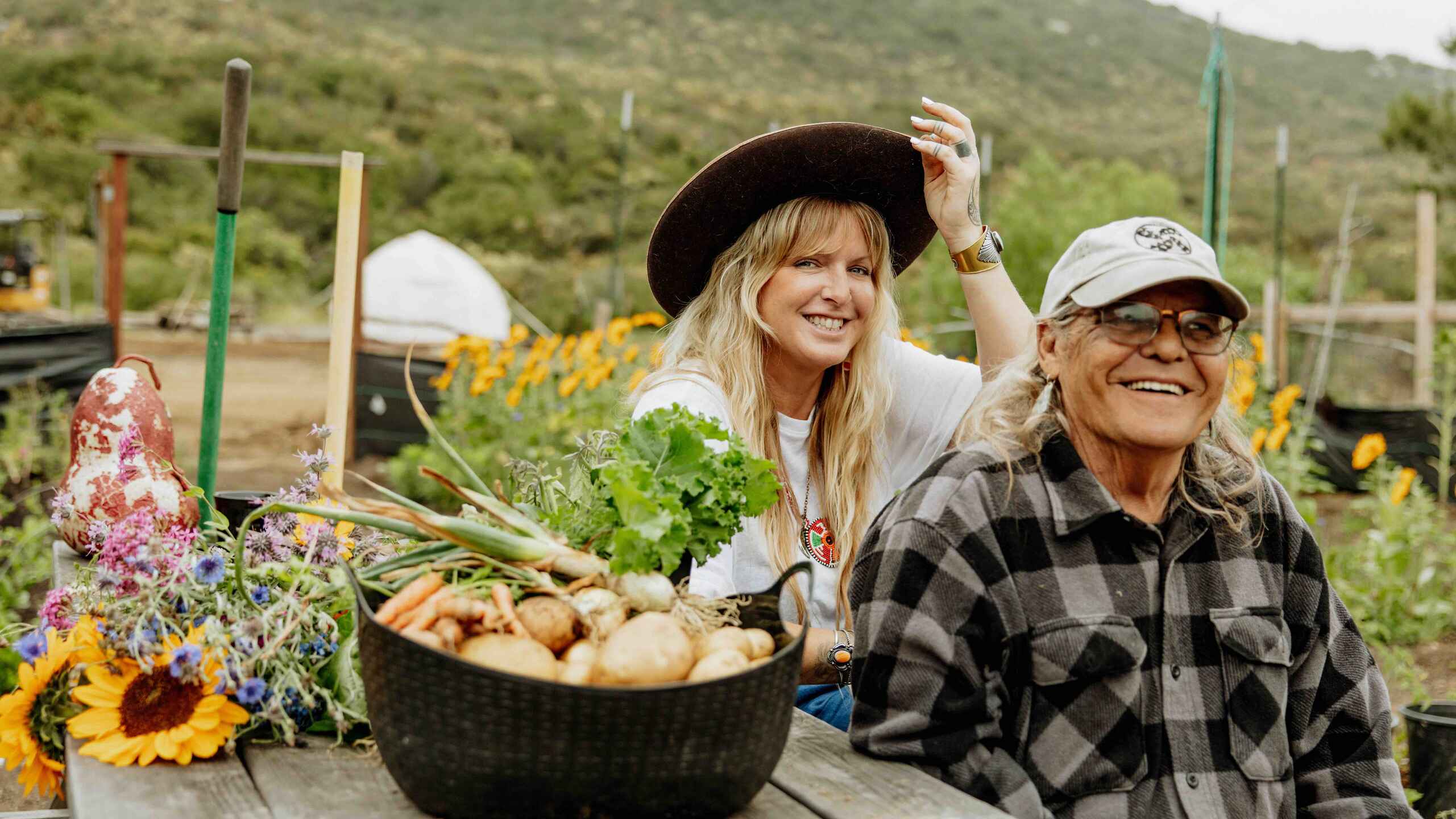 Two farmers display harvested vegetables and flowers from their regenerative garden with mountains in the background