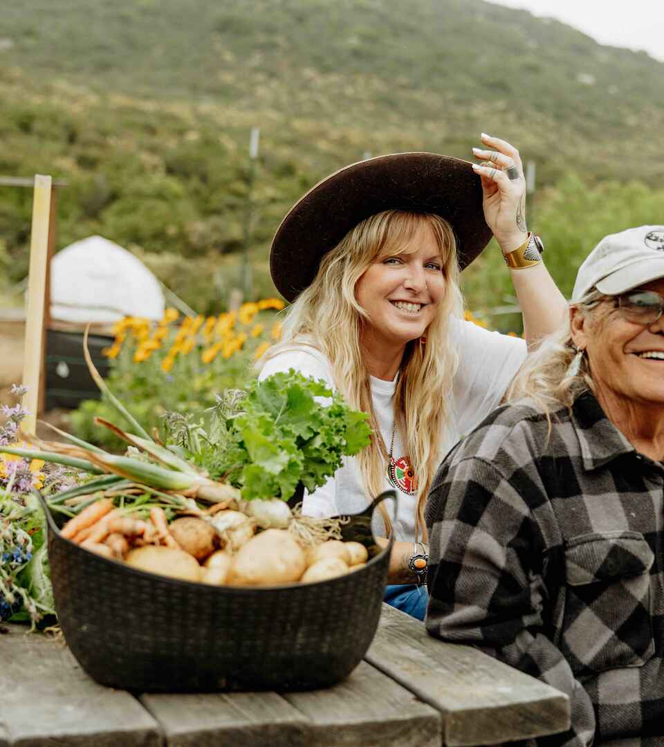 Two farmers display harvested vegetables and flowers from their regenerative garden with mountains in the background