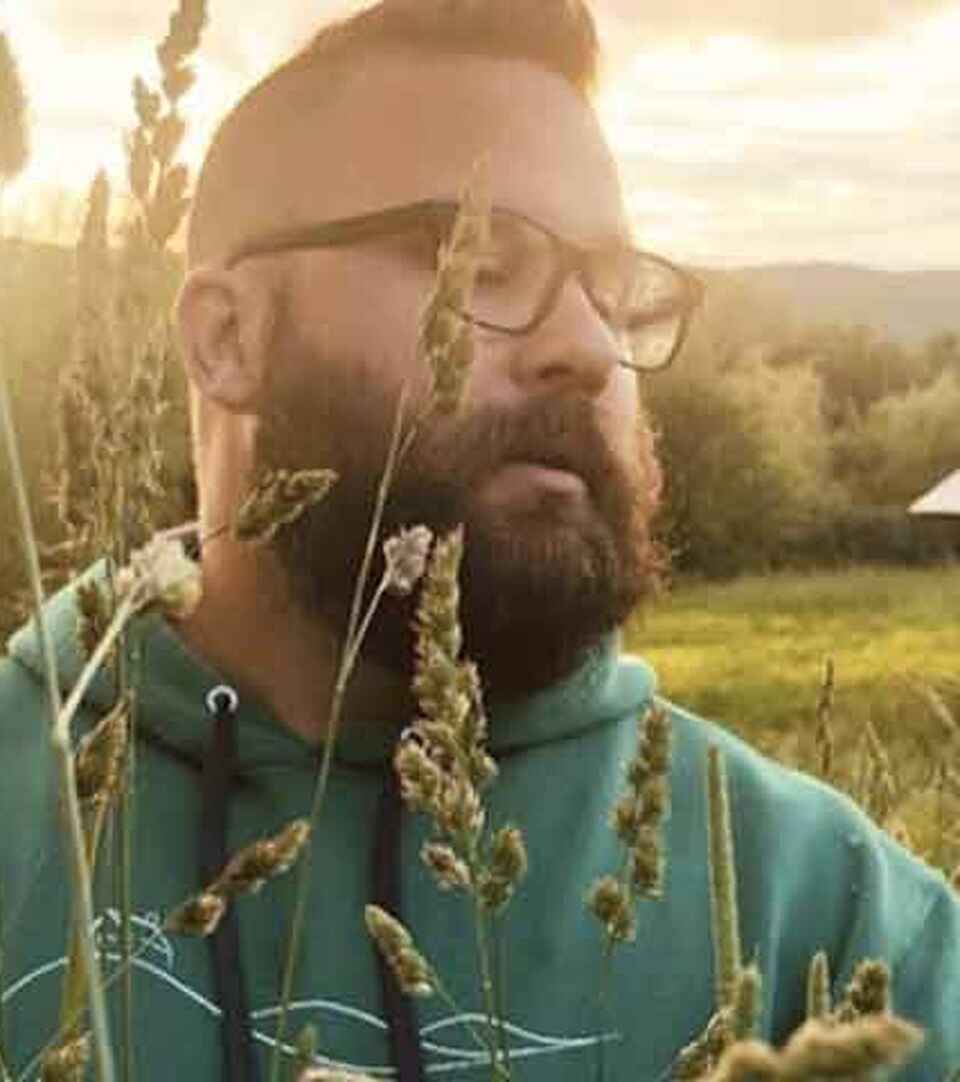 Agricultor con barba y gafas examinando cultivos de sorgo en un campo con paneles solares y edificios agr&iacute;colas al fondo