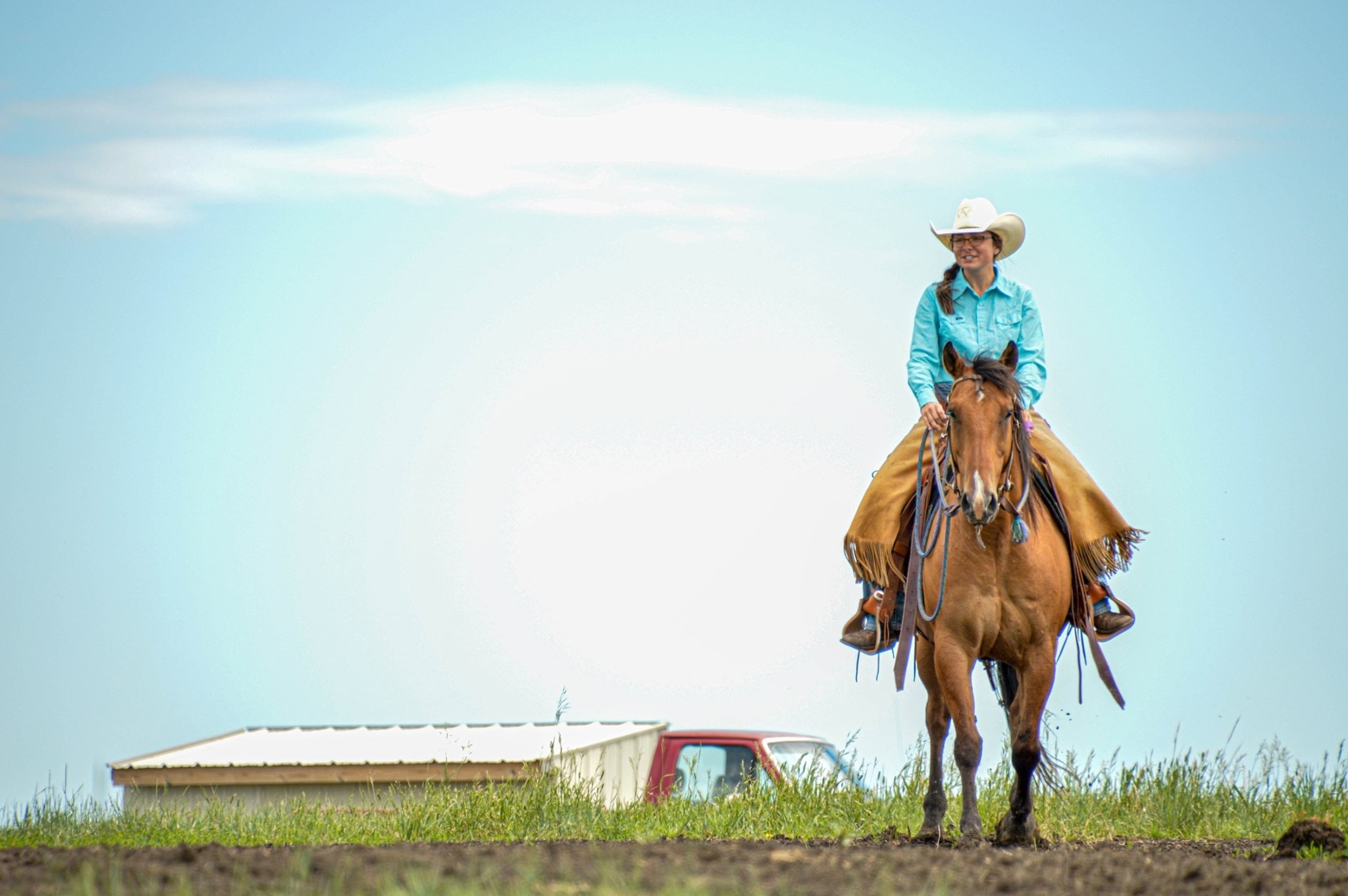 Rancher on horseback in turquoise shirt and white cowboy hat overlooking prairie land with farm equipment