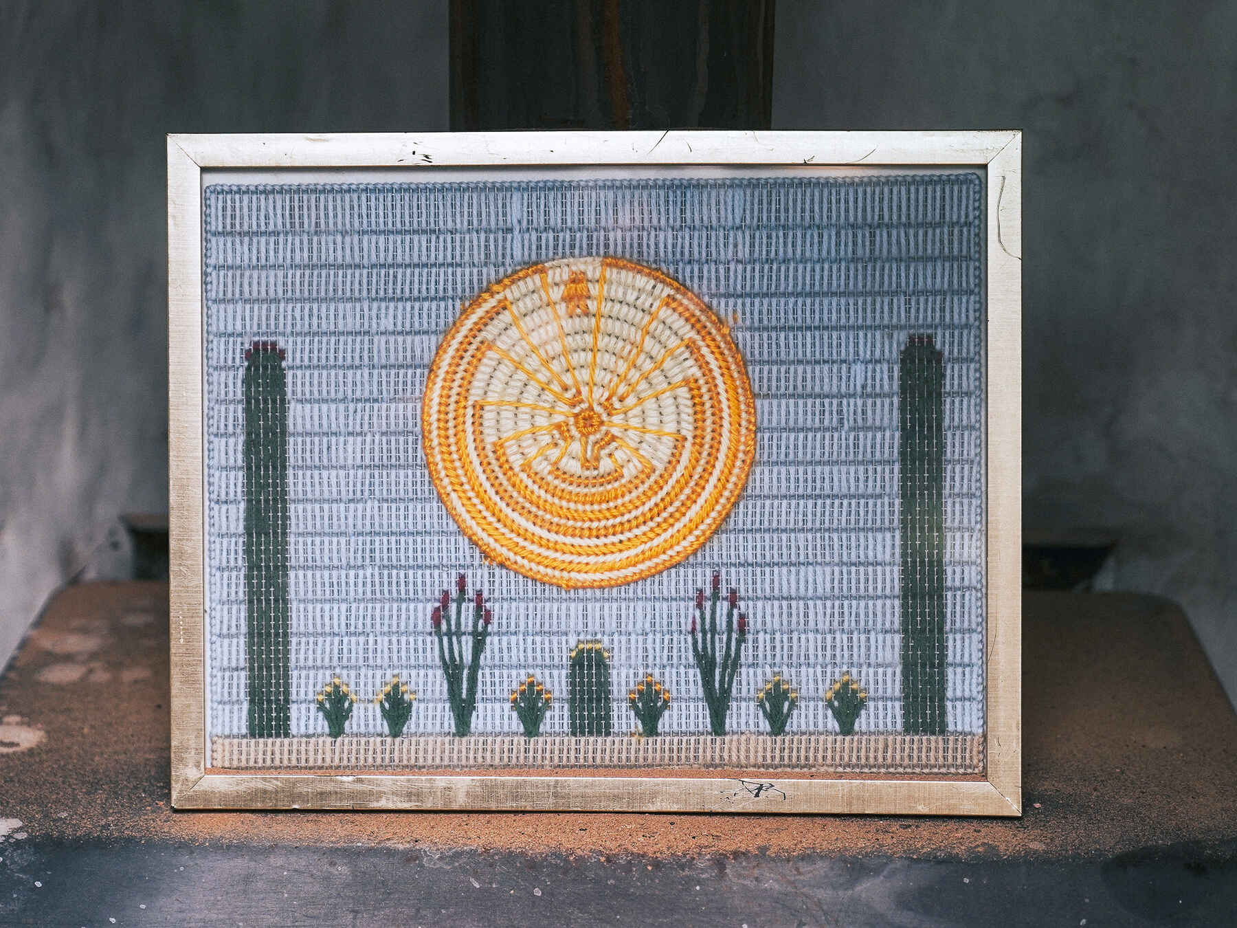 Cross-stitch artwork depicting a Hopi desert landscape with a golden sun, saguaro cacti, and smaller desert plants against a blue sky