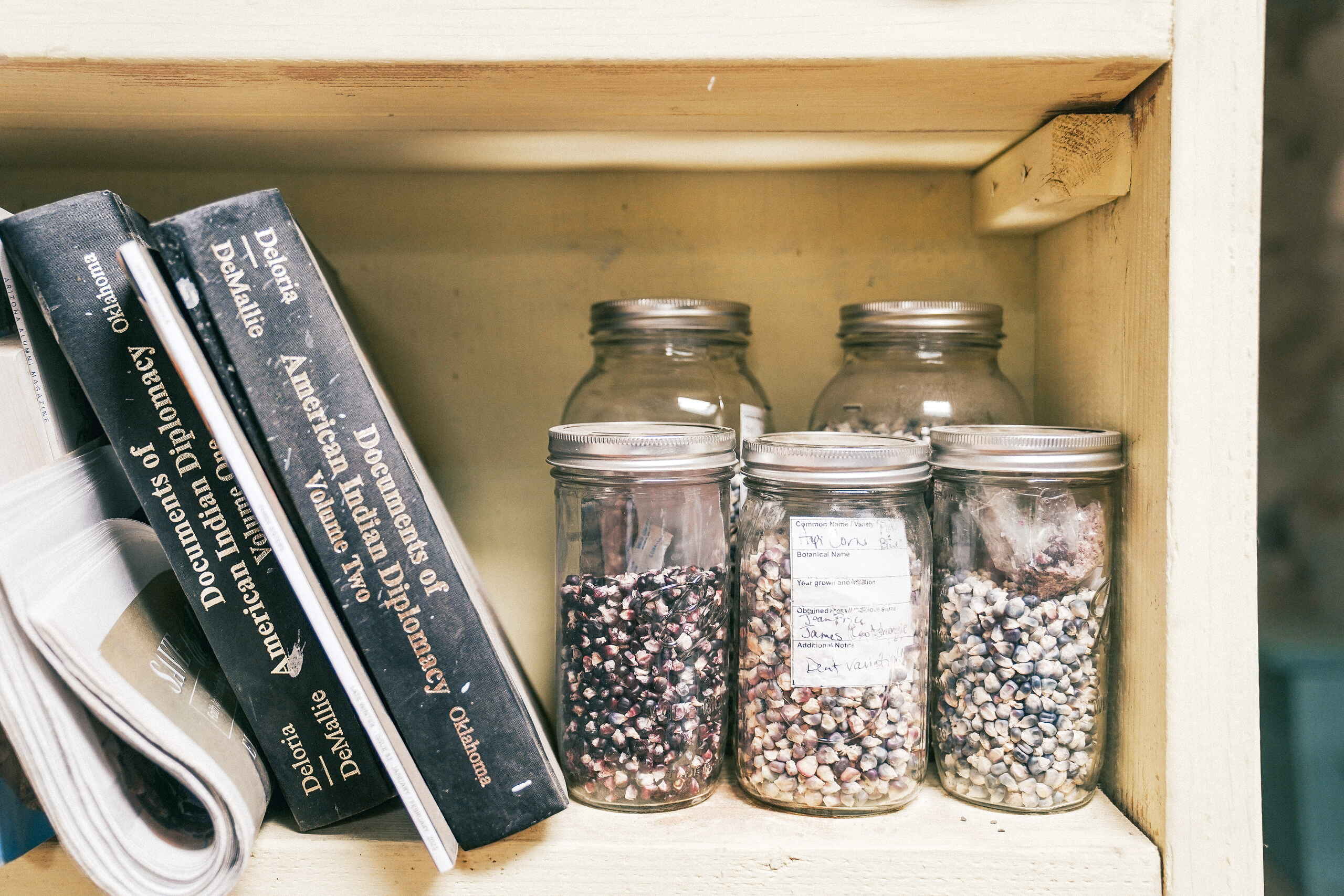 Wooden shelf with two books about indigenous agriculture next to glass jars containing different varieties of corn seeds and legumes