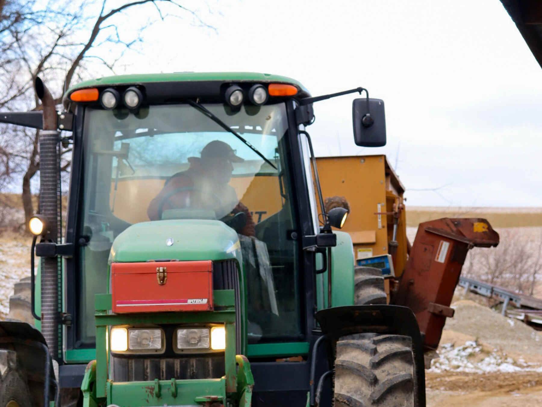 Farmer operating a green John Deere tractor in a farm yard with equipment and bare trees visible in the background