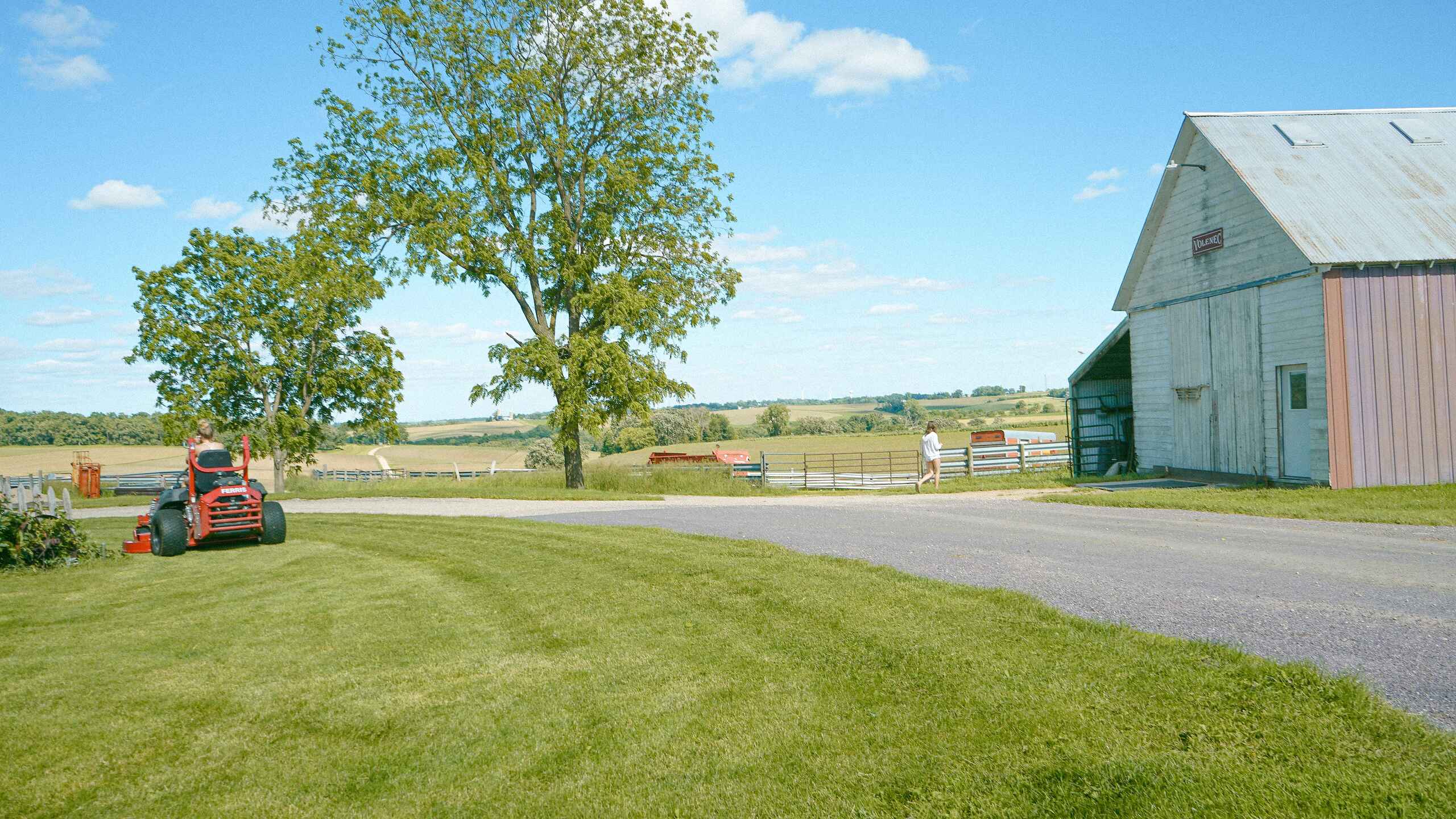 Red tractor mowing grass near a farmhouse and barn on a rural property