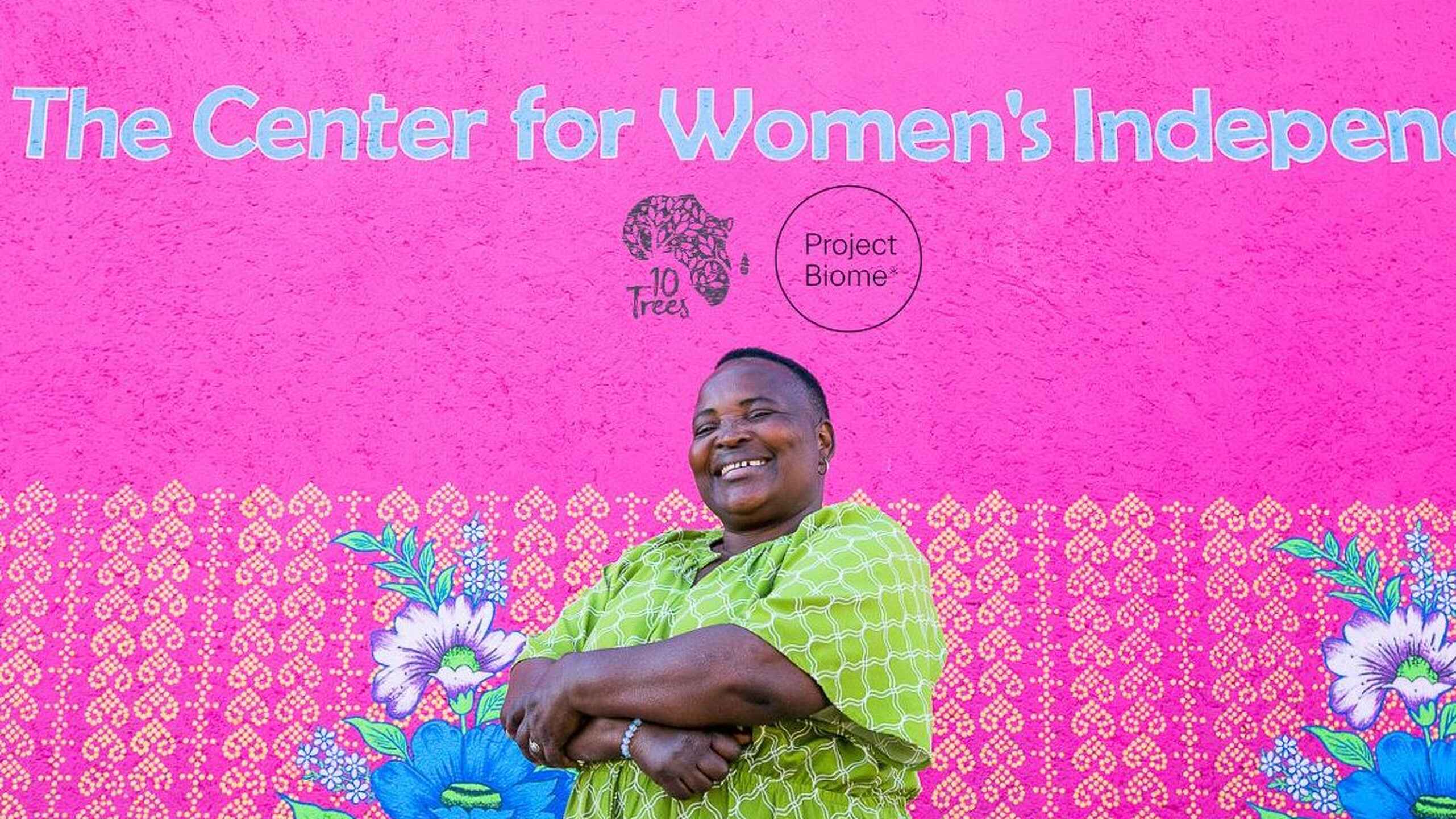 Woman in a lime green patterned dress smiling in front of a bright pink mural for The Center for Women's Independence and 10 Trees Project Biome