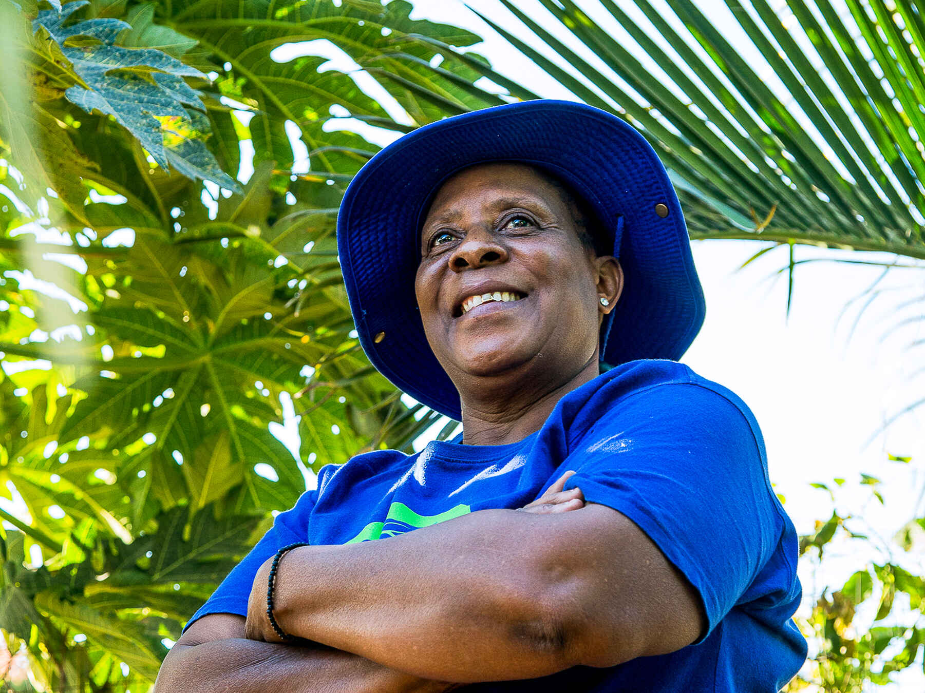 Woman in blue shirt and hat smiling while standing among lush green plants
