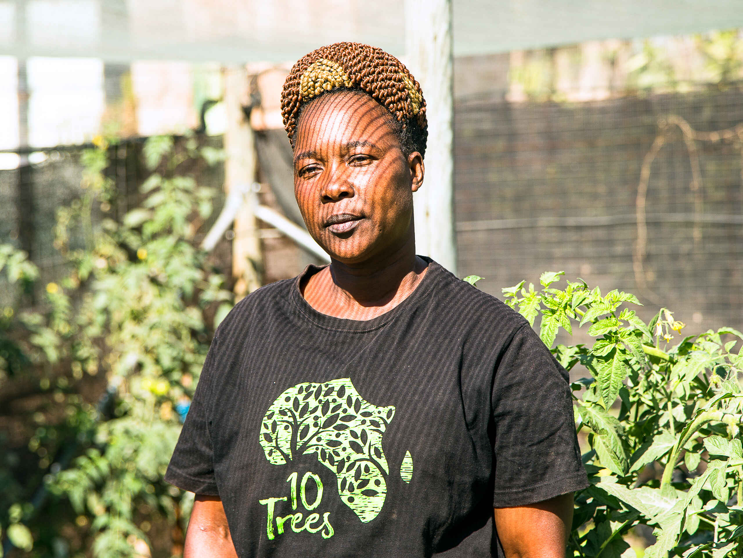 Winnie Shongwe stands in a garden wearing a 10 Trees t-shirt, surrounded by growing plants and vegetables