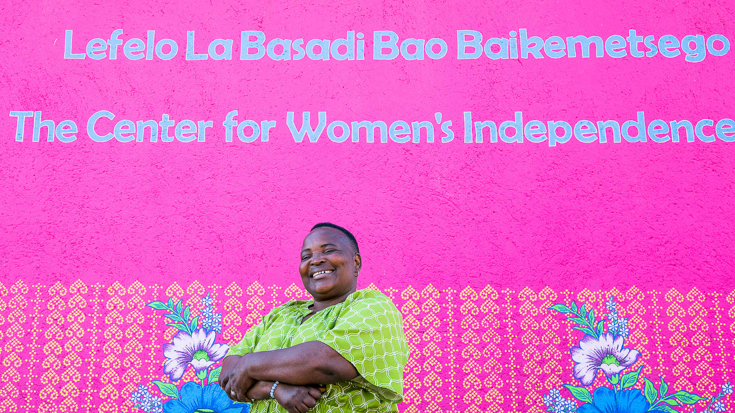 Woman in a lime green patterned dress smiling in front of a bright pink wall with text about The Center for Women's Independence