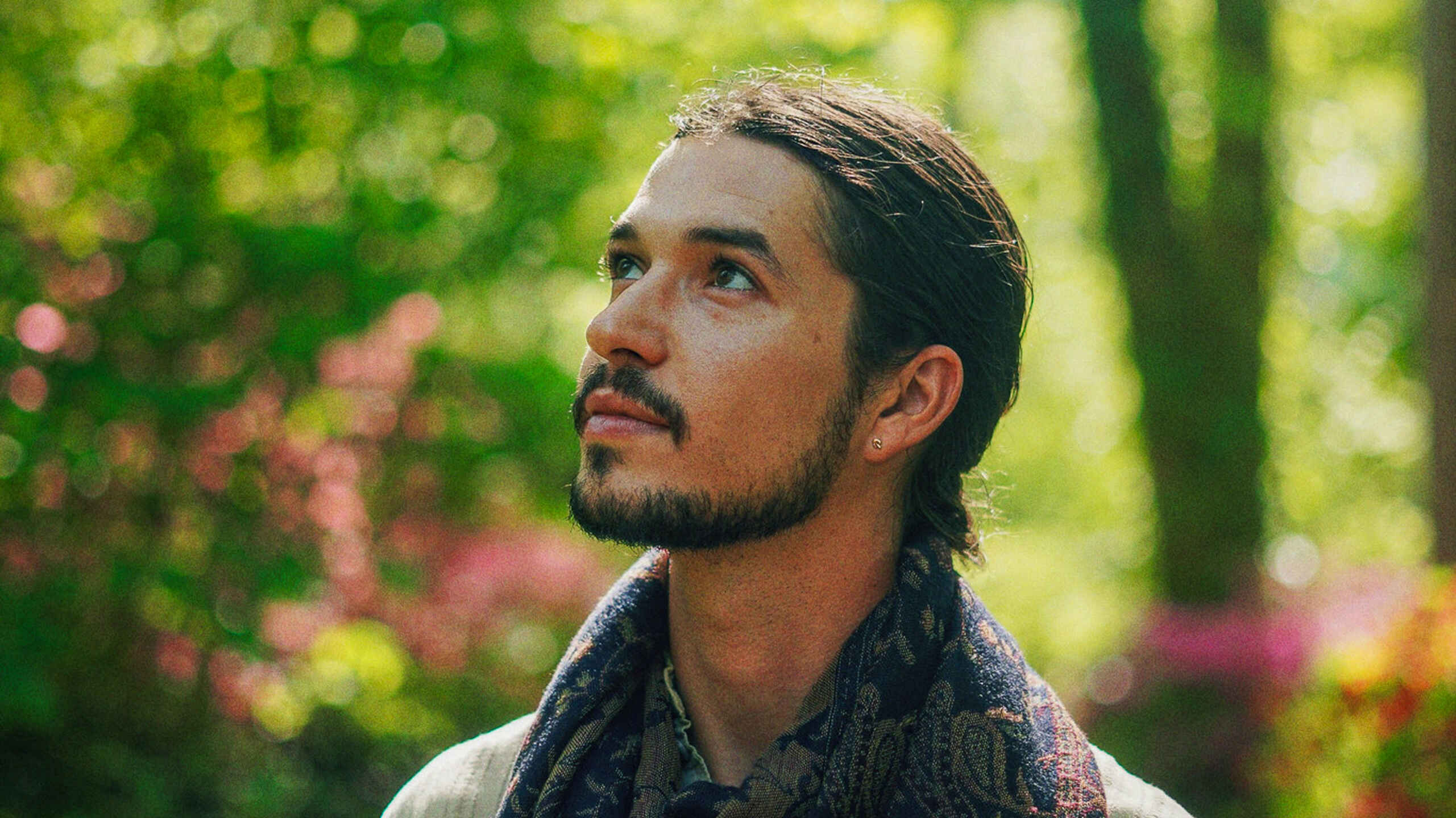 Bodhi Vasilopoulos stands in a garden wearing a light-colored shirt and patterned scarf, looking upward among blooming flowers and tall trees.