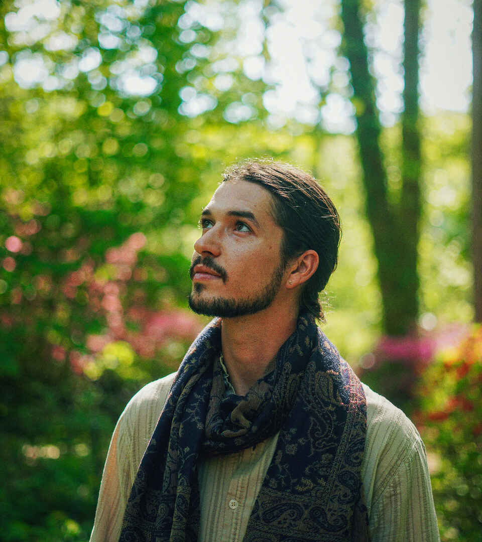 Bodhi Vasilopoulos stands in a garden wearing a light-colored shirt and patterned scarf, looking upward among blooming flowers and tall trees.