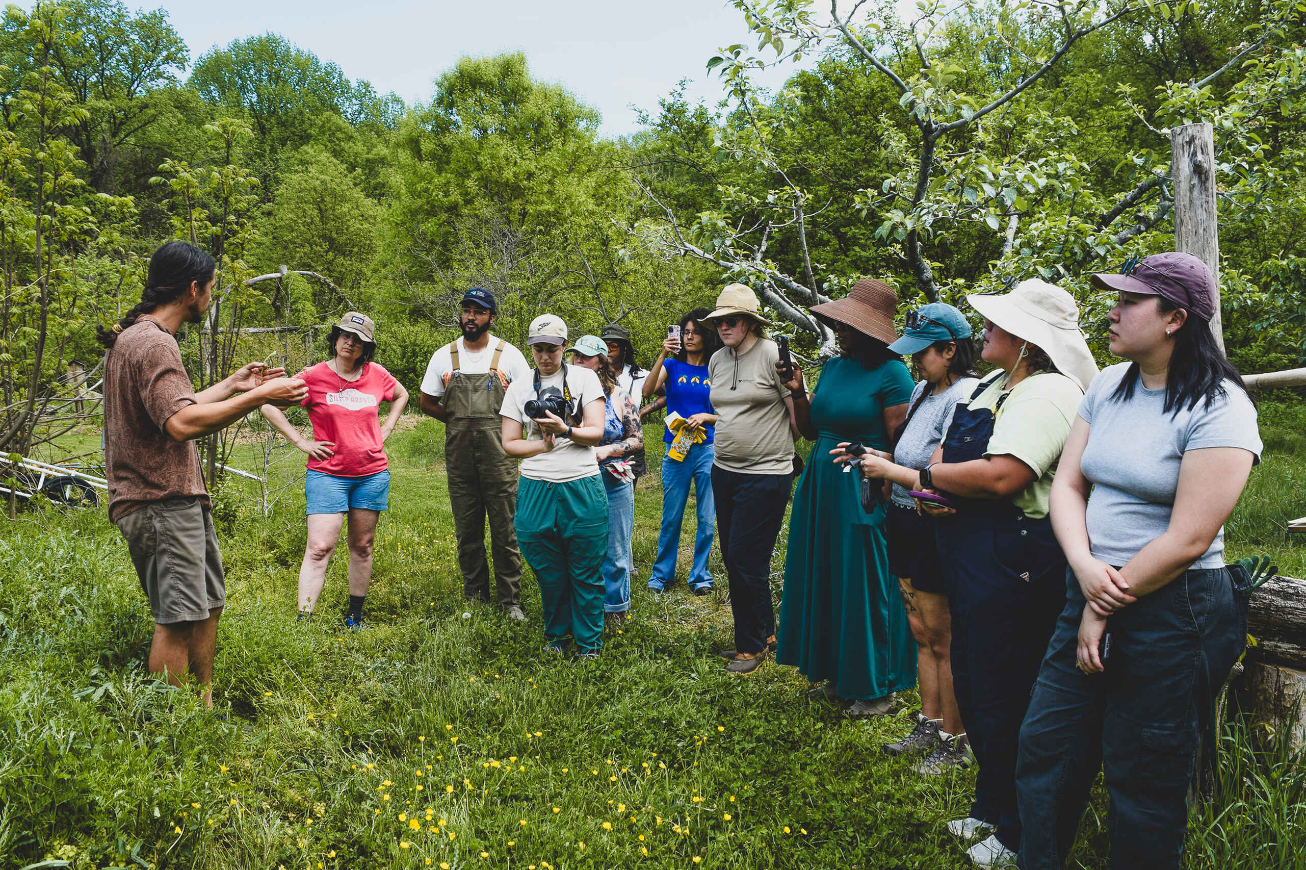 A foraging instructor leads an outdoor educational workshop with a diverse group of participants gathered in a grassy woodland area