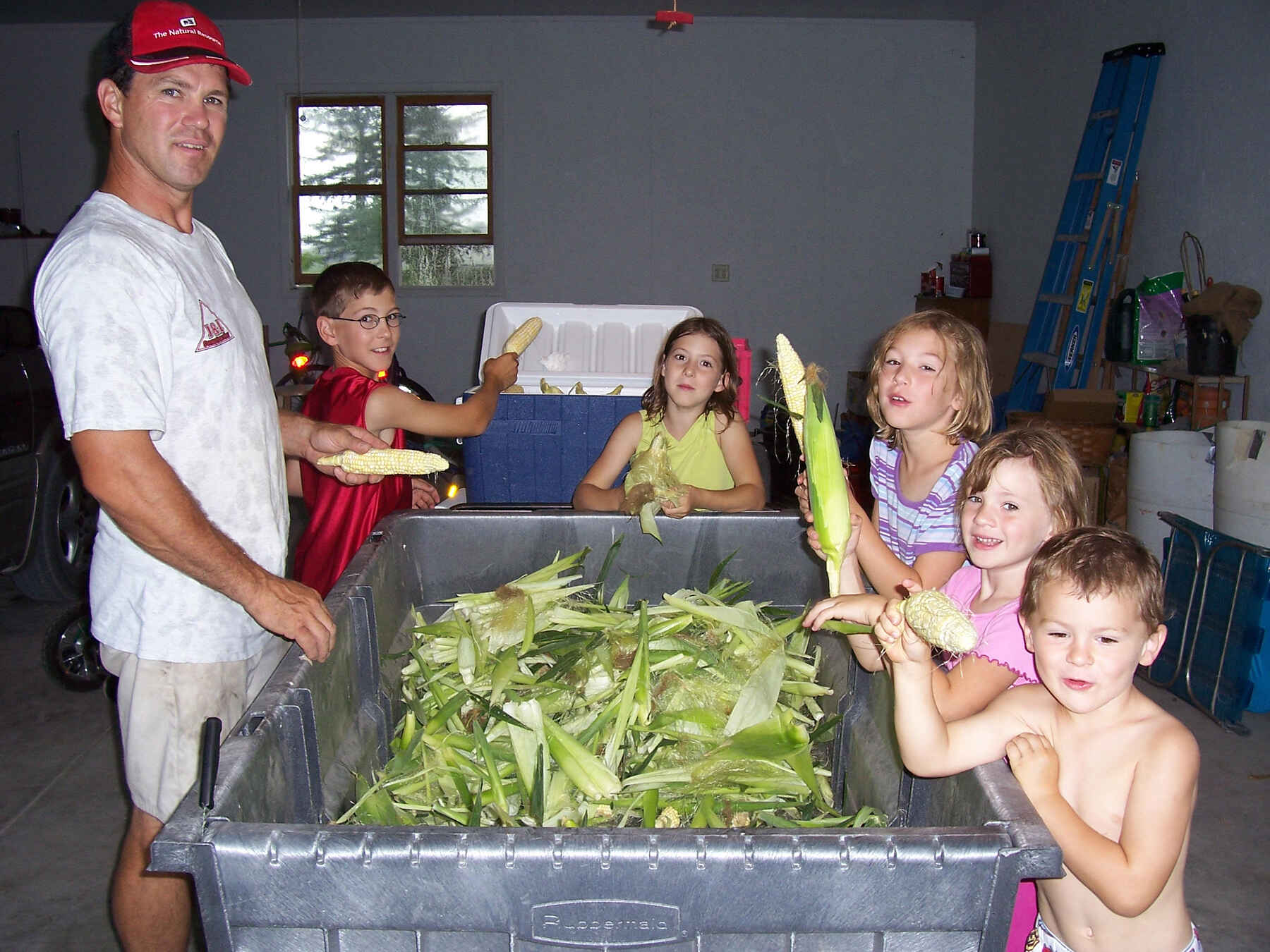 An adult and six children stand around a large bin filled with fresh corn ears, holding and displaying the harvest