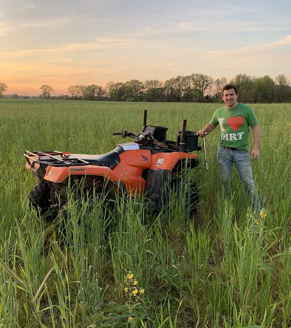 Mitchell Hora stands next to an orange ATV in a green grain field at sunset