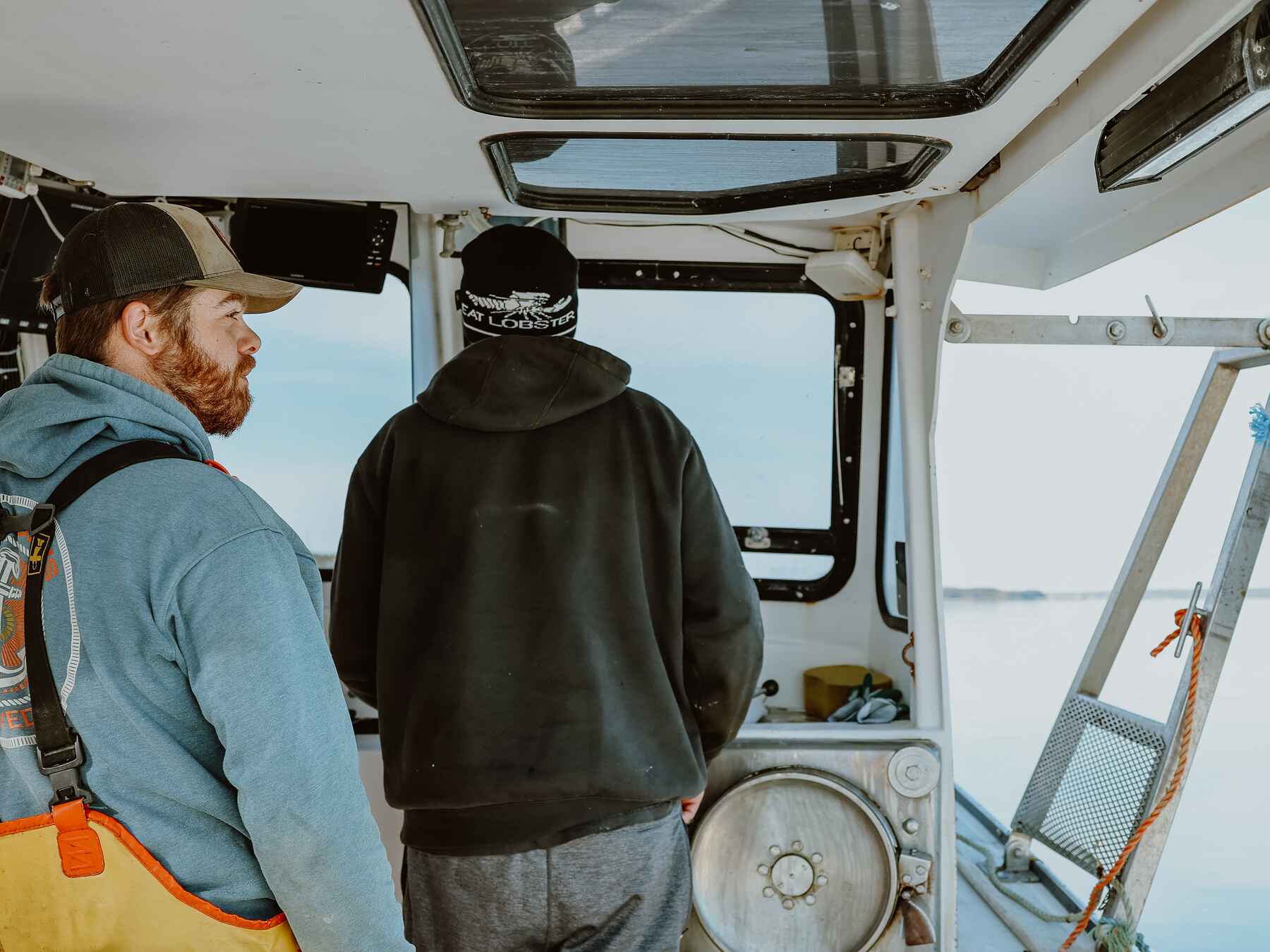 Two people in safety gear stand inside a fishing vessel cabin looking out at calm waters