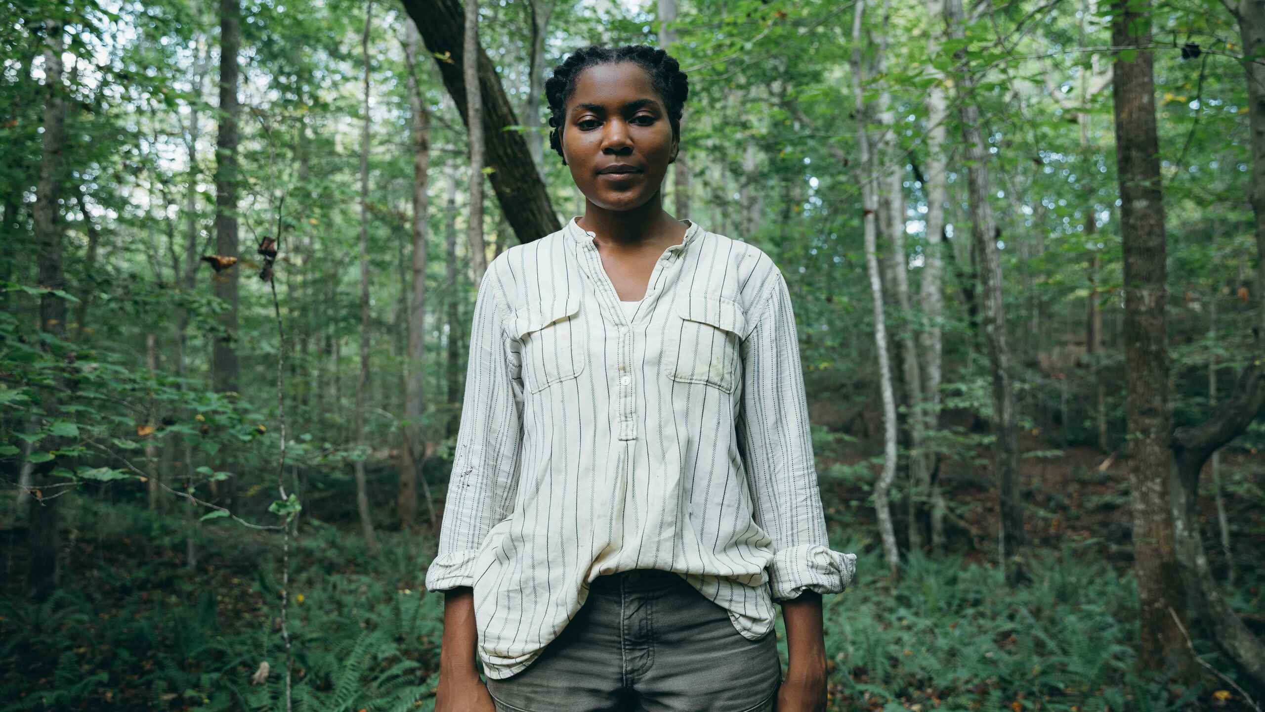 Woman standing in a forest wearing a light pinstriped shirt and dark pants