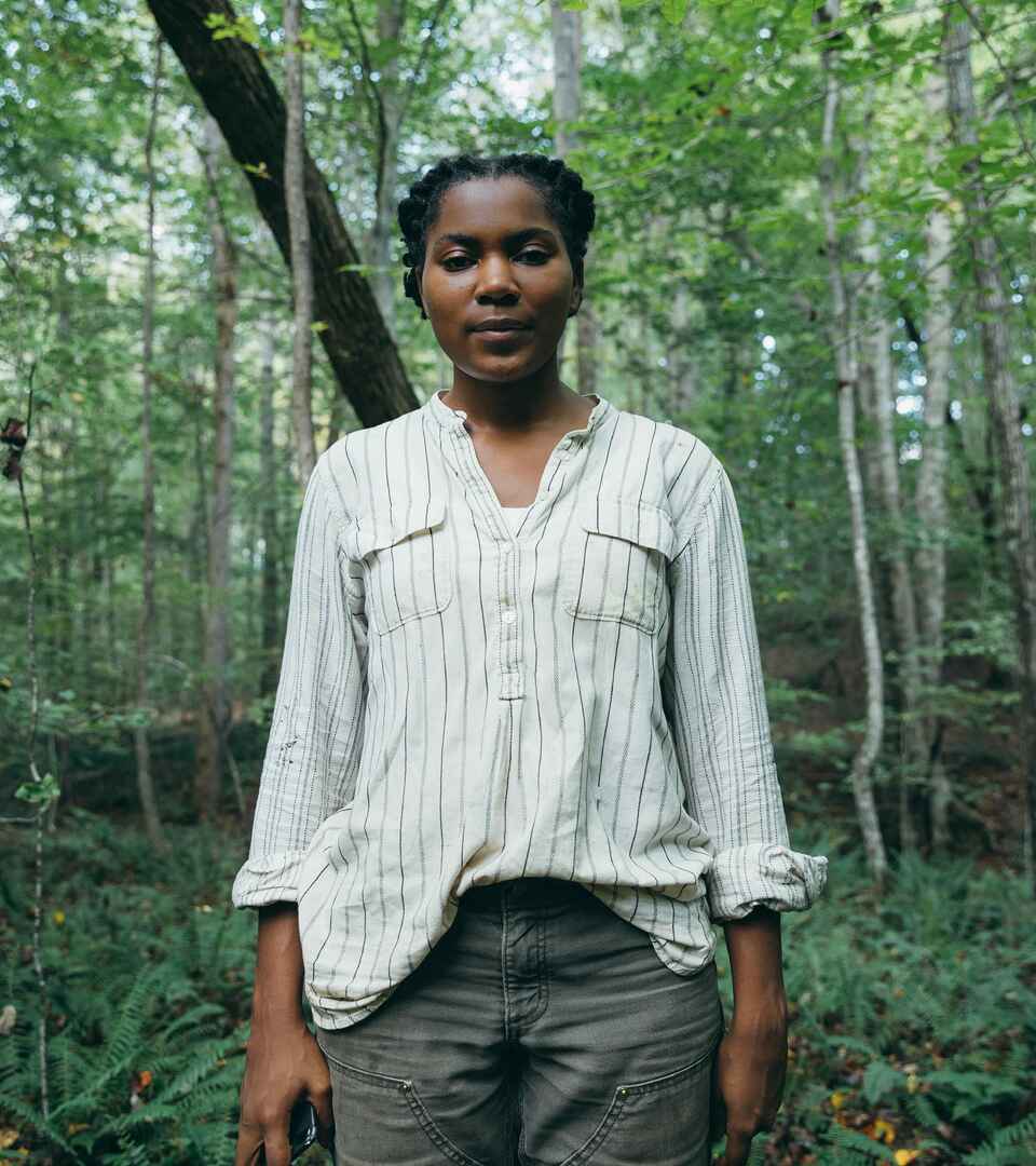 Woman standing in a forest wearing a light pinstriped shirt and dark pants