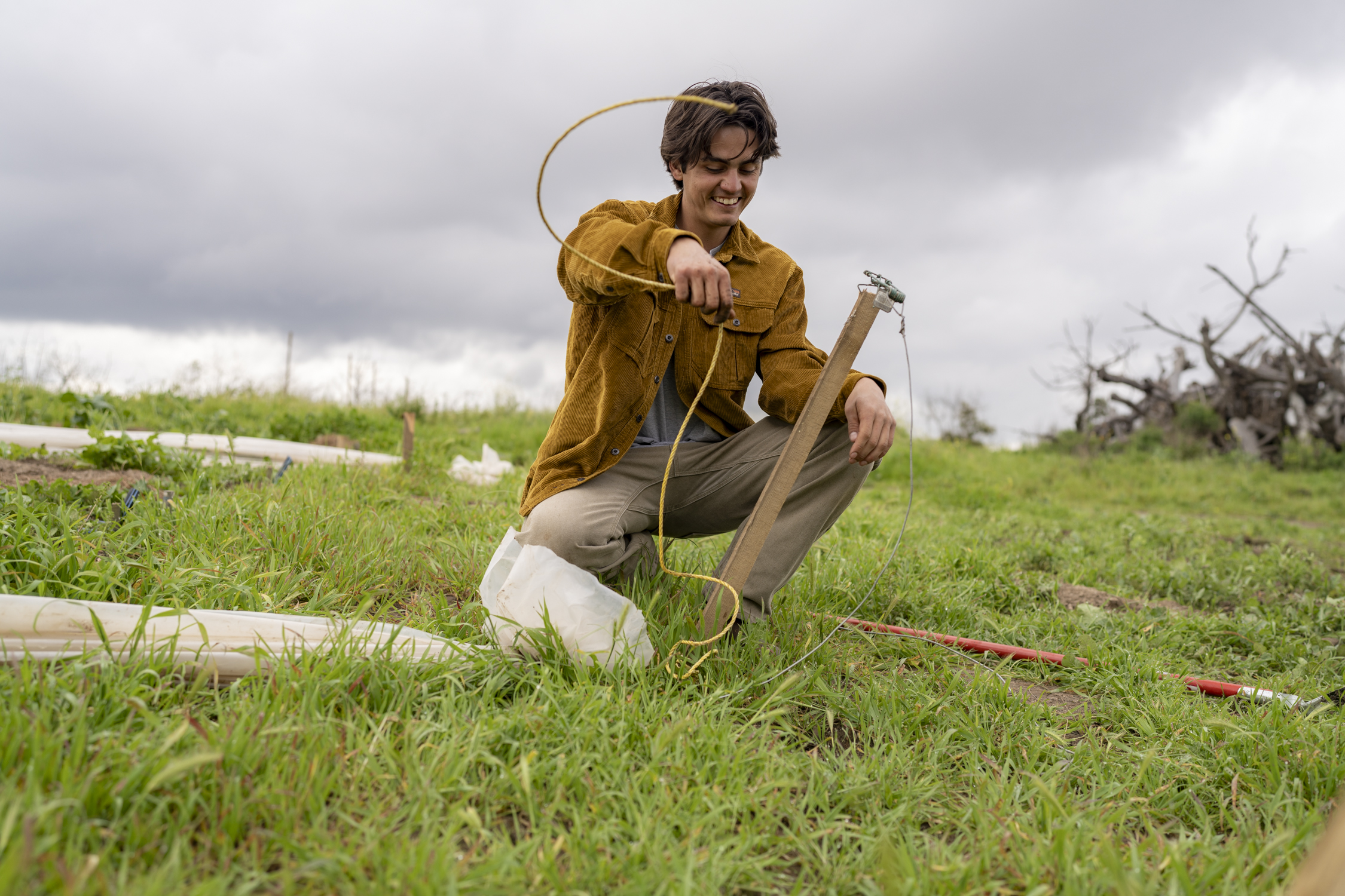 A farmer crouches in an irrigated field while holding a shovel and coiled hose, inspecting the irrigation system