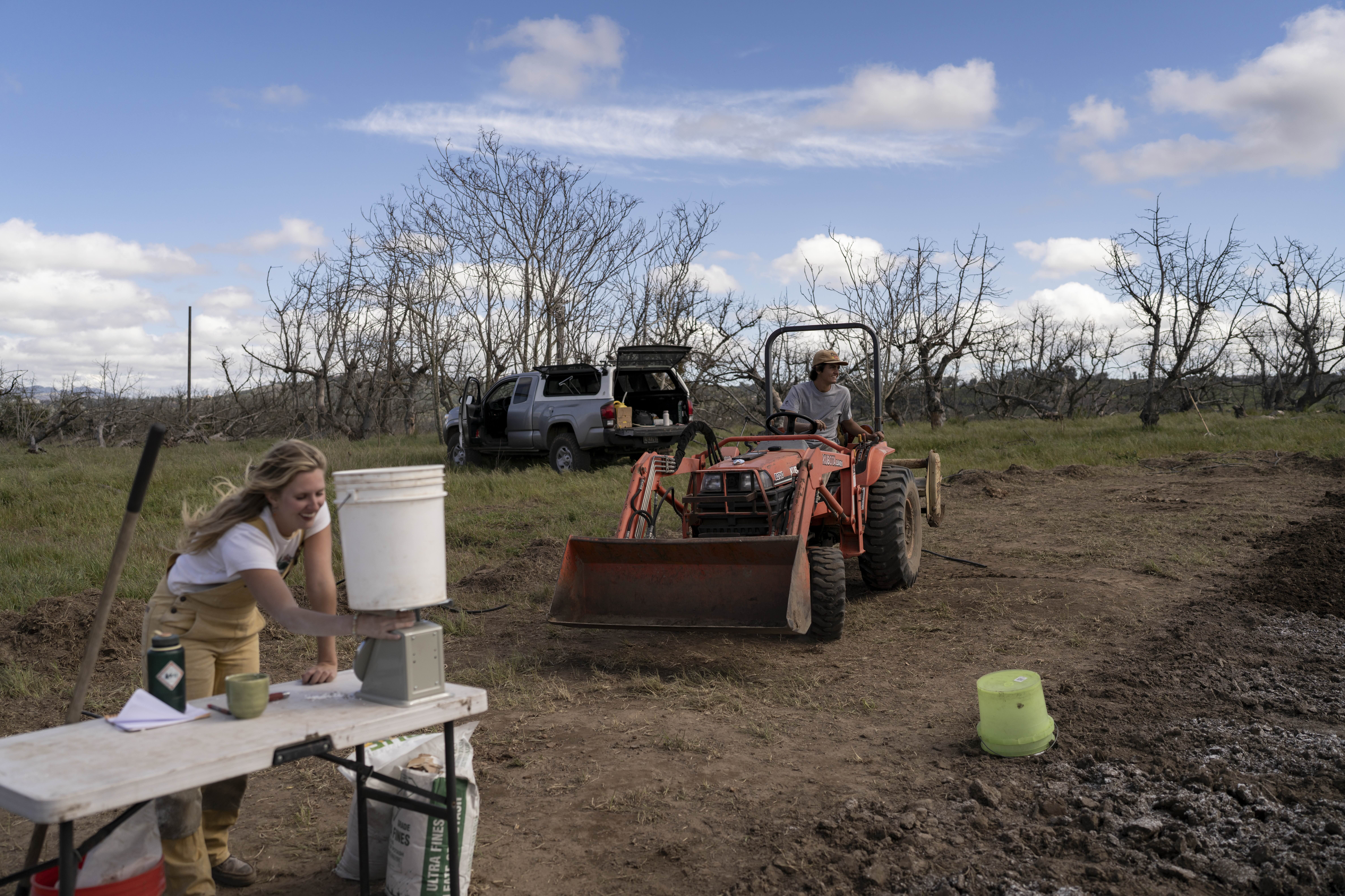 Woman operating water testing equipment while farmer operates tractor with blade in regenerative agriculture field