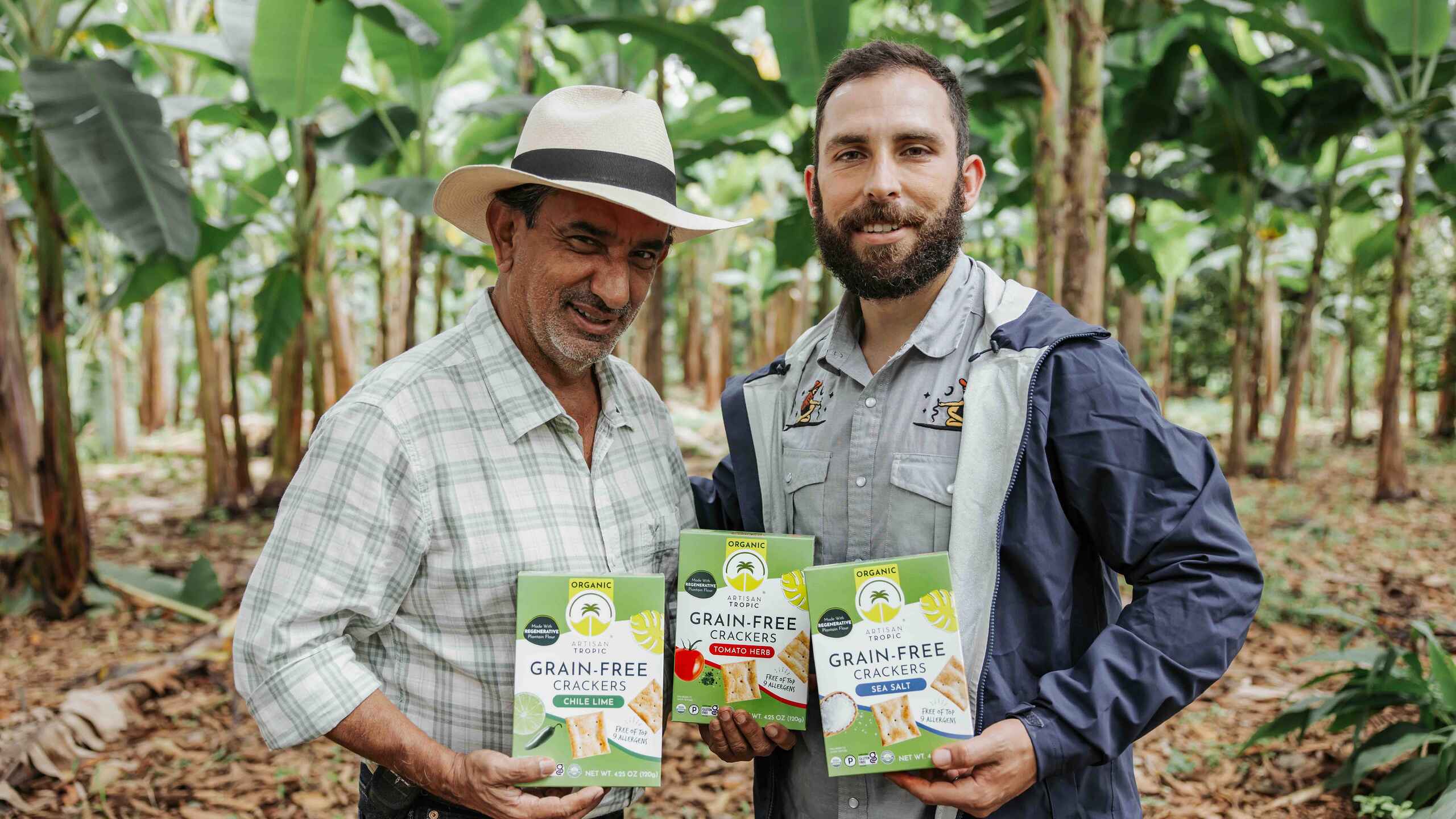 Two men holding boxes of organic grain-free crackers in a banana plantation