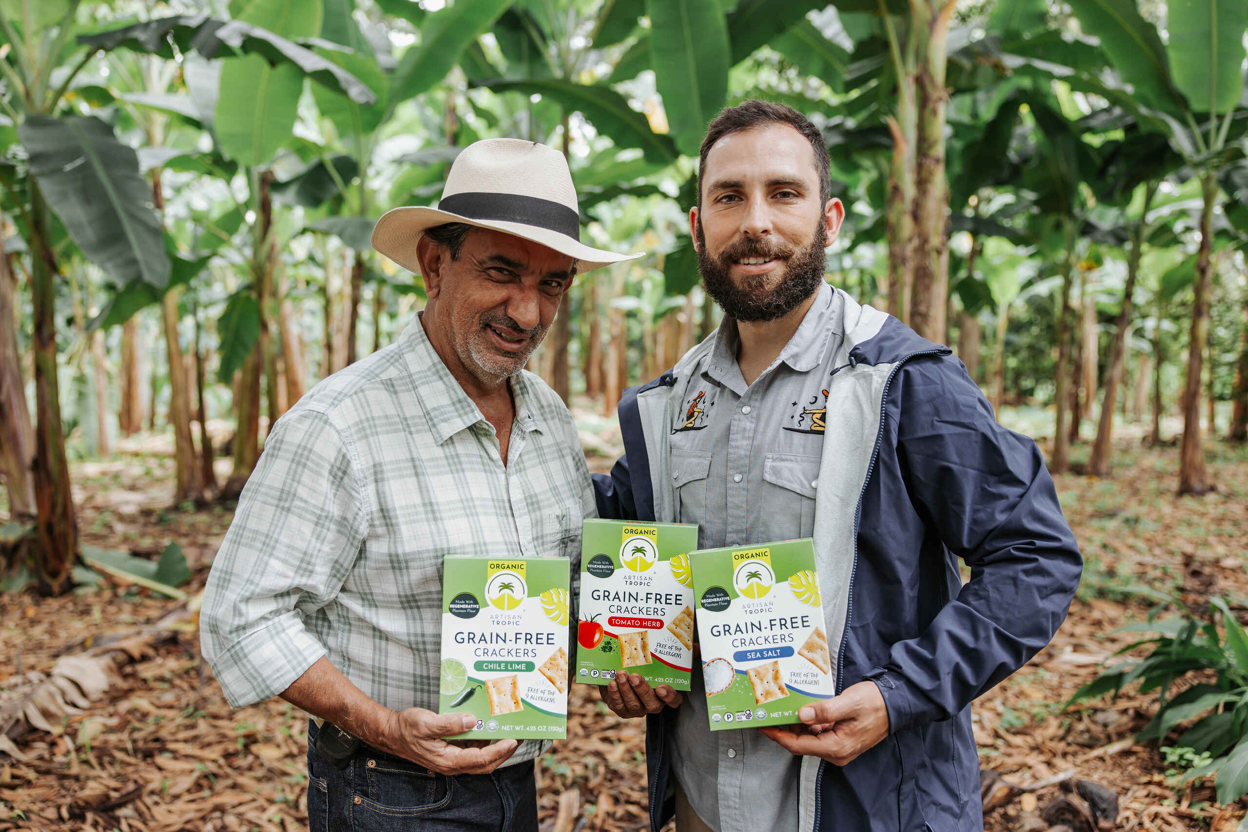 Two men holding grain-free cracker boxes in a banana plantation