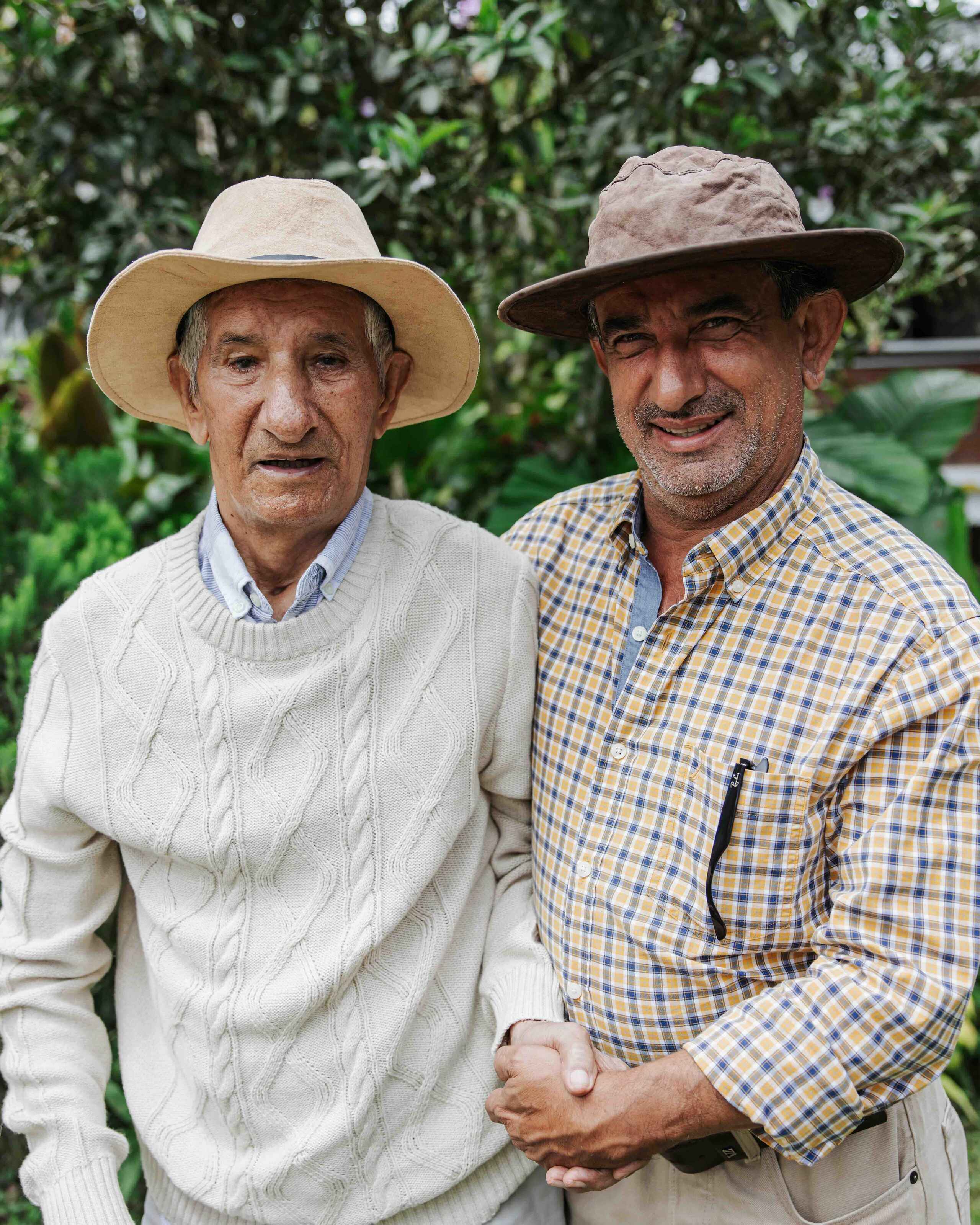 Two farmers shake hands while wearing cowboy hats in front of lush green vegetation