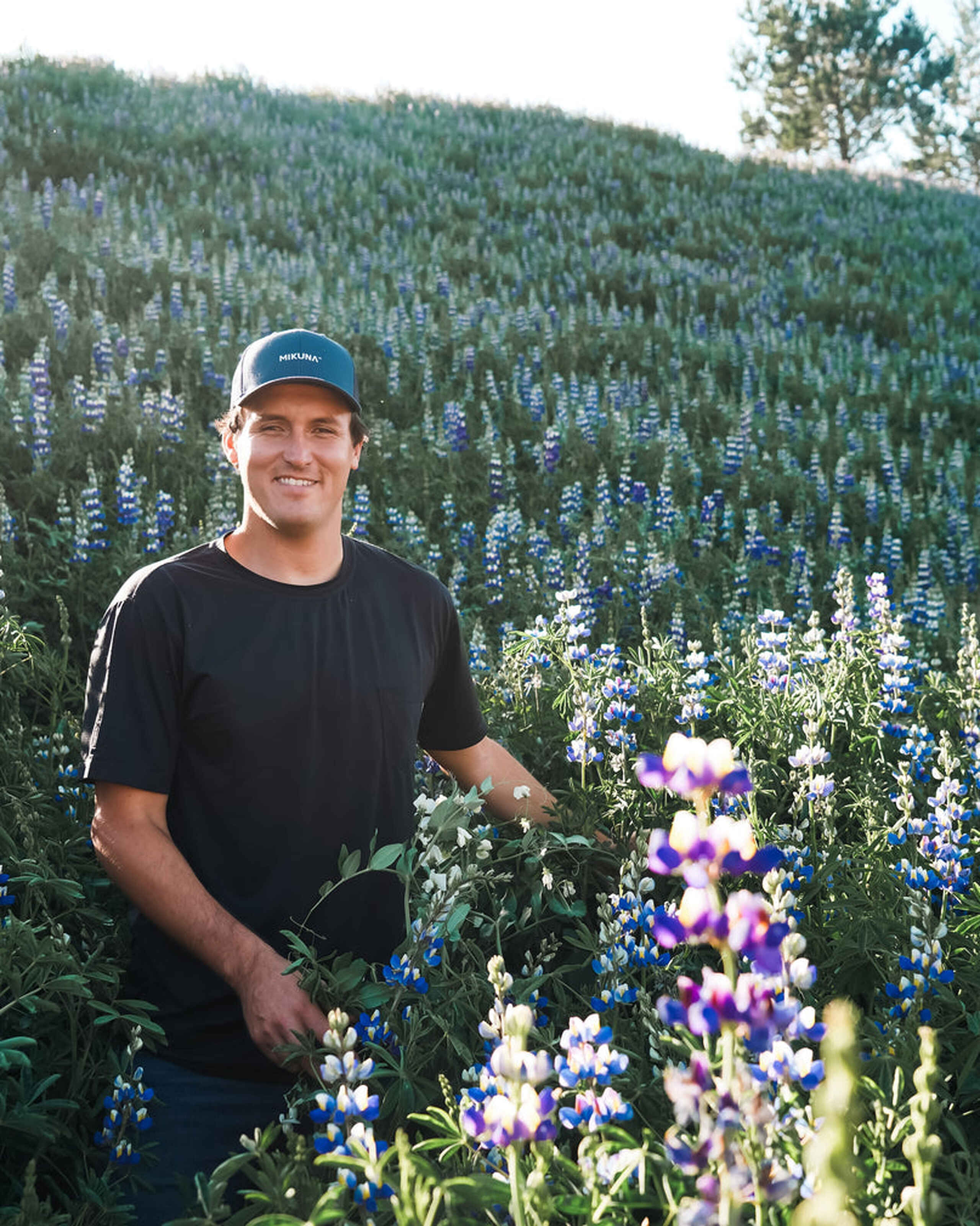 Ricky Mikuna de pie en un campo de flores de lupino p&uacute;rpura y blanco con monta&ntilde;as boscosas al fondo