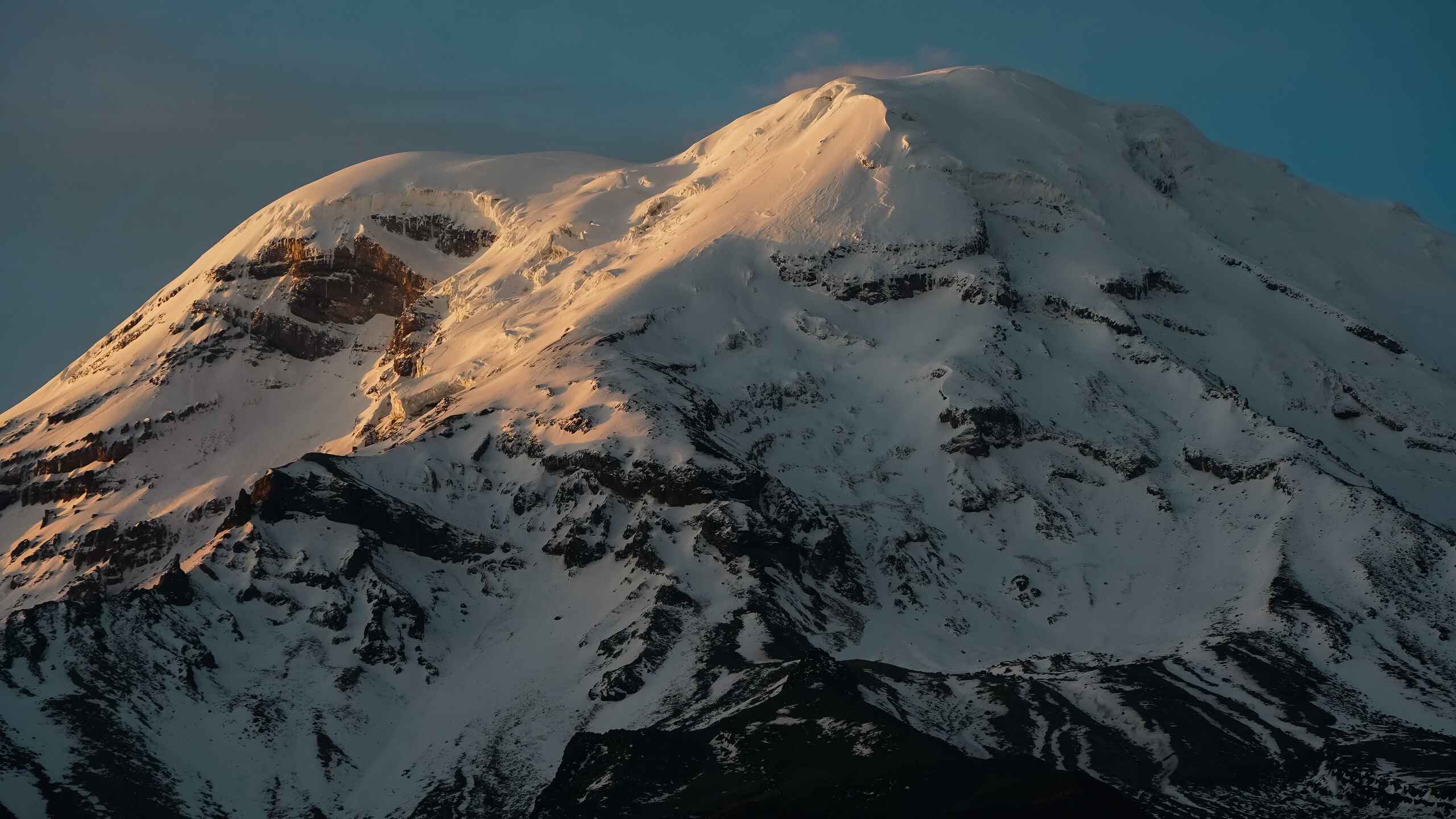 Snow-covered volcanic mountain peak illuminated by golden sunlight against a clear blue sky