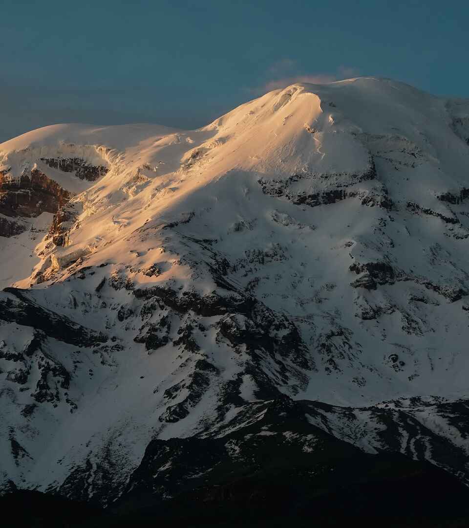Snow-covered volcanic mountain peak illuminated by golden sunlight against a clear blue sky