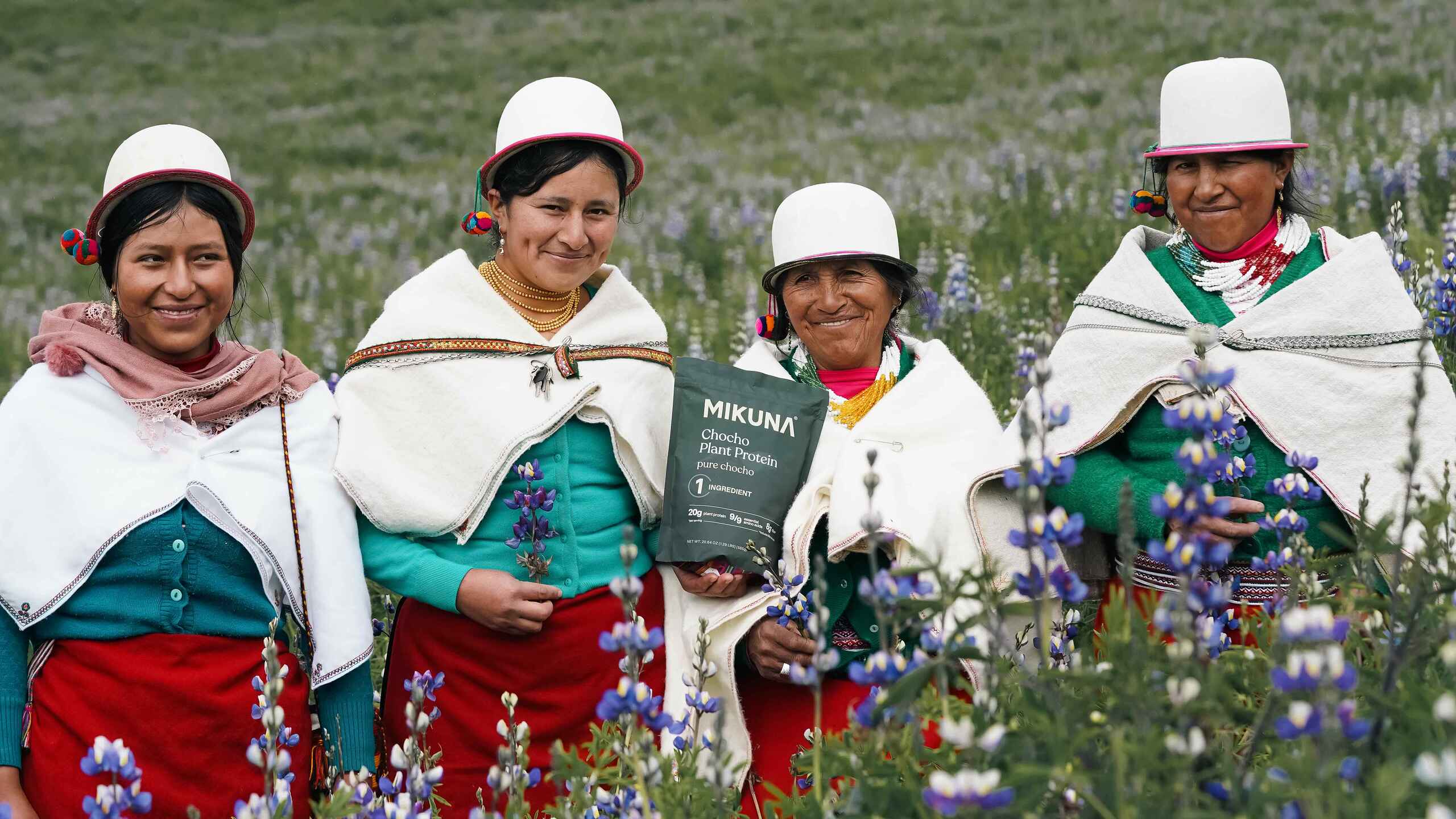 Four Andean women in traditional dress holding a package of Mikuna chocolate in a field of blue lupine flowers