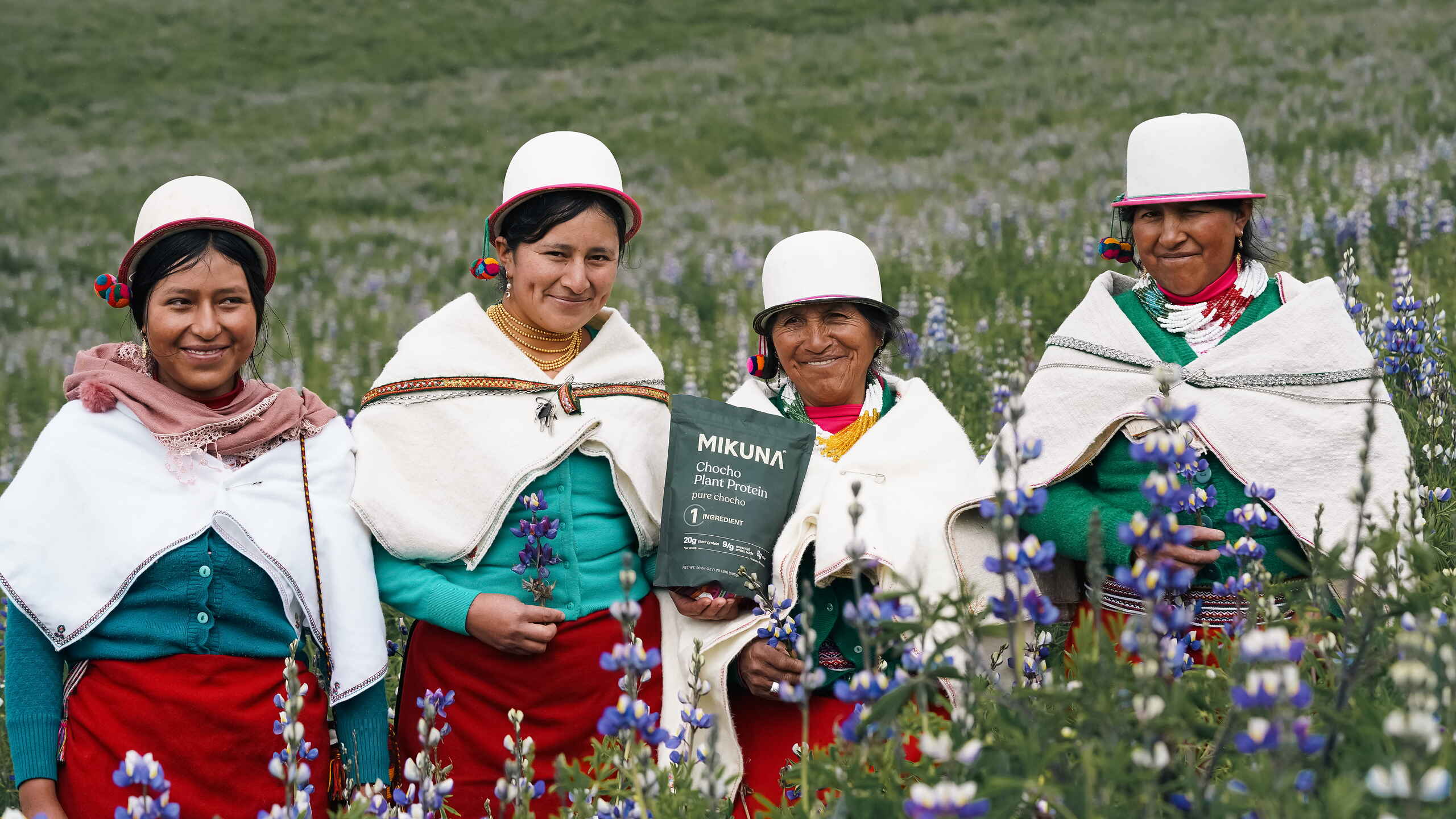 Cuatro mujeres ind&iacute;genas andinas con vestimenta tradicional sosteniendo un producto de chocolate MIKUNA en un campo de flores lupino