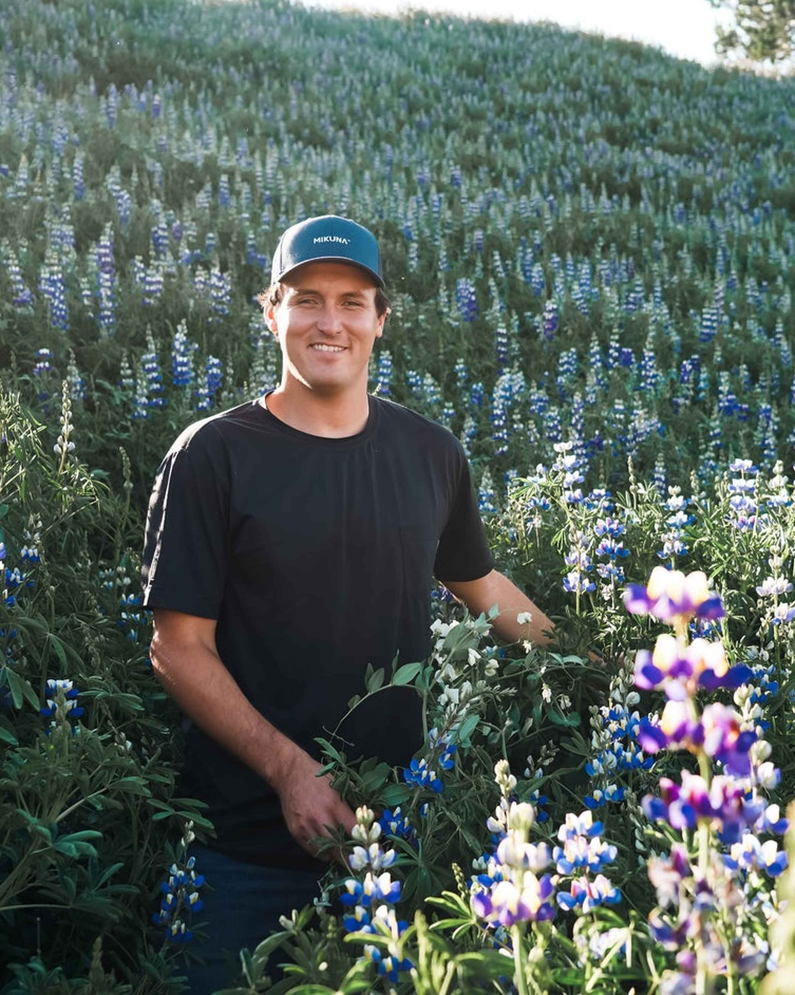 Man in a black shirt and cap smiling in a field of blue and purple lupine flowers with forested mountains in the background