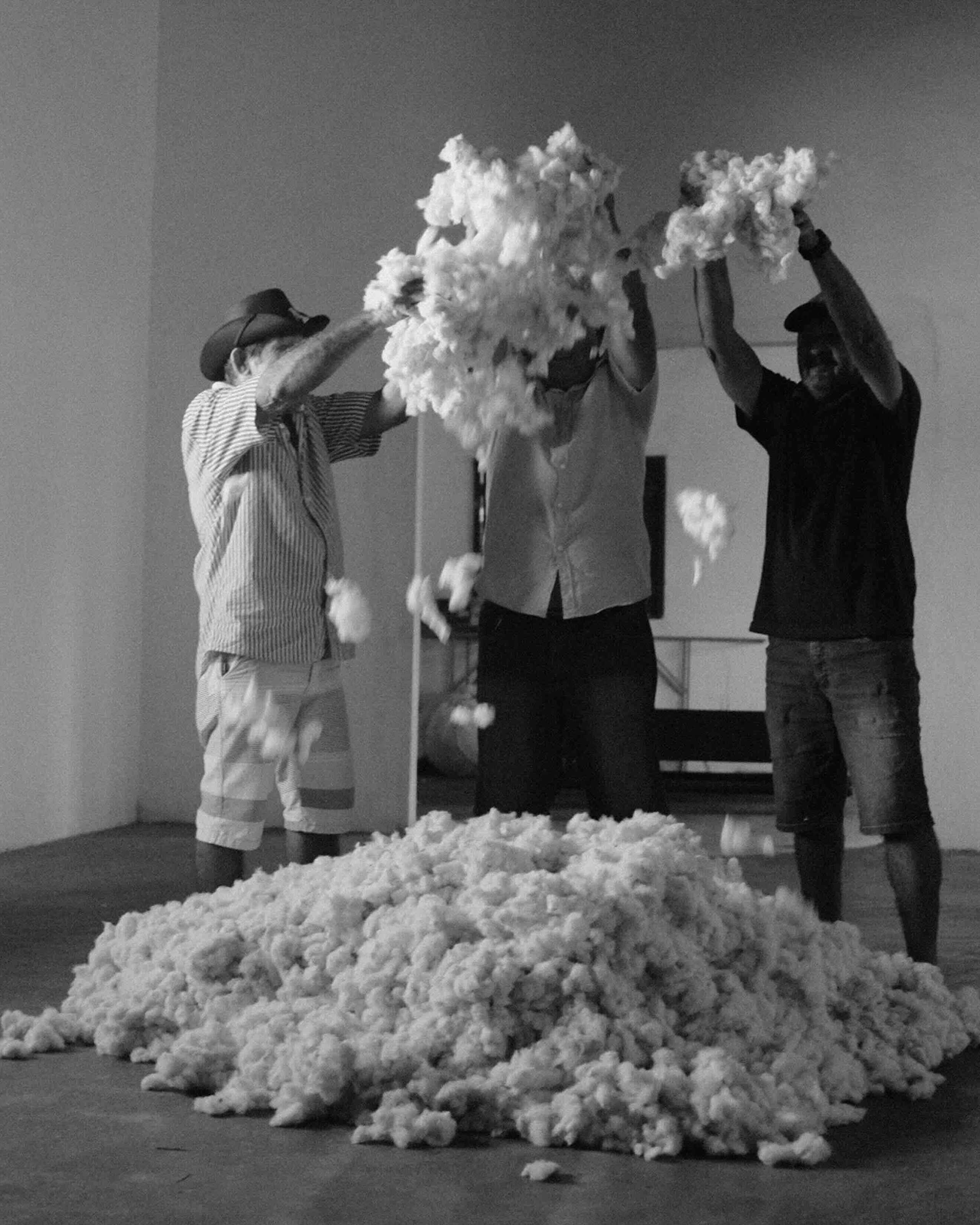 Three people toss cotton or wool fibers into the air inside a gallery space, creating clouds of white material