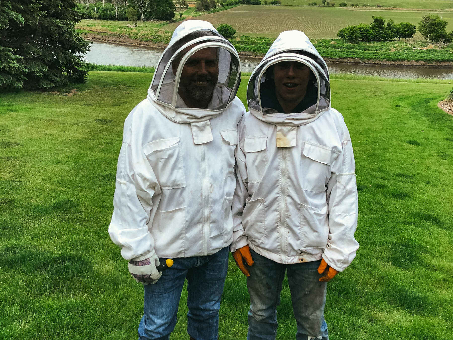 Two beekeepers in protective suits stand in a green pasture with farmland and a river in the background
