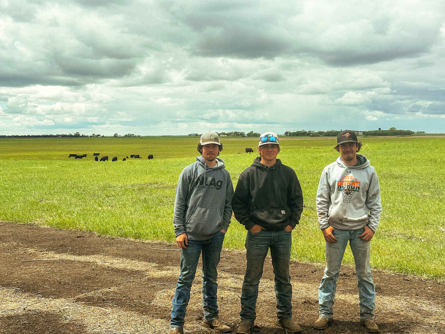 Three young men stand together in a dirt area overlooking a vast green pasture with cattle grazing in the distance under a cloudy sky