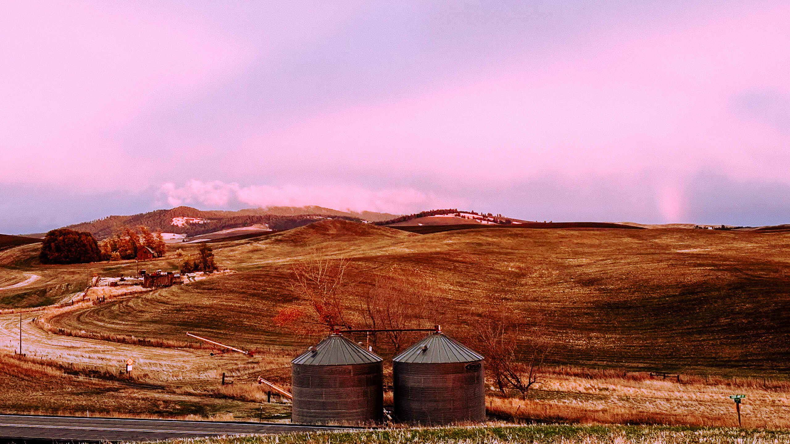 Two grain storage silos sit in a rolling agricultural landscape at sunset