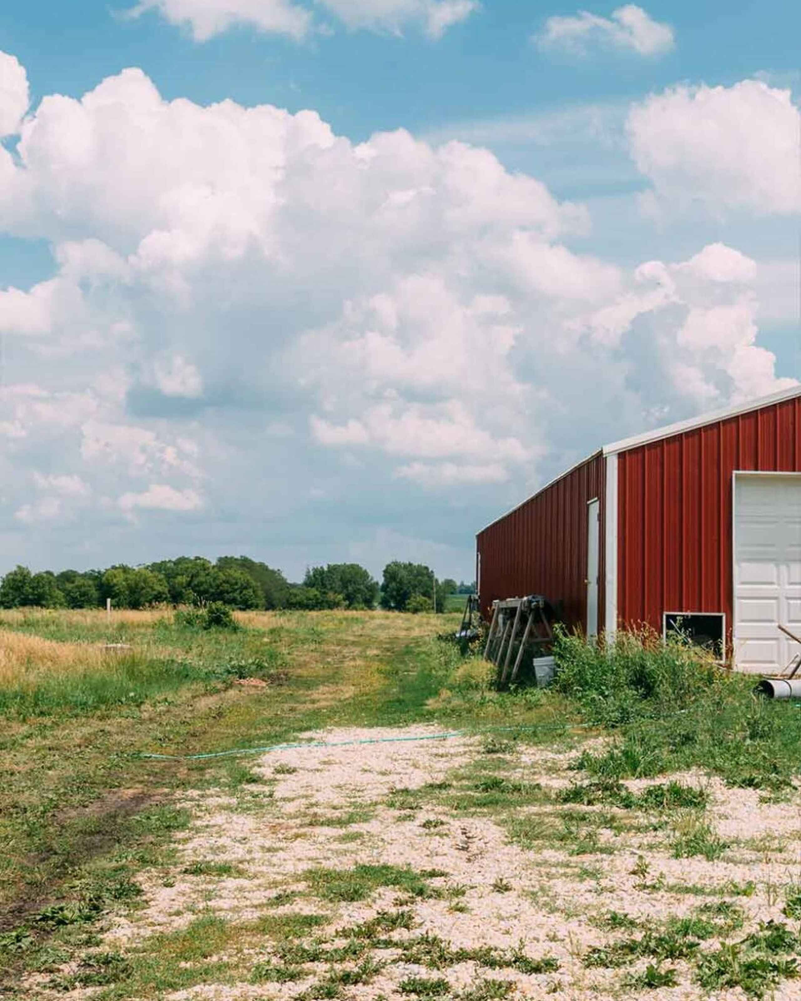 Red metal barn with white door standing in sparse grassland under blue sky with white clouds