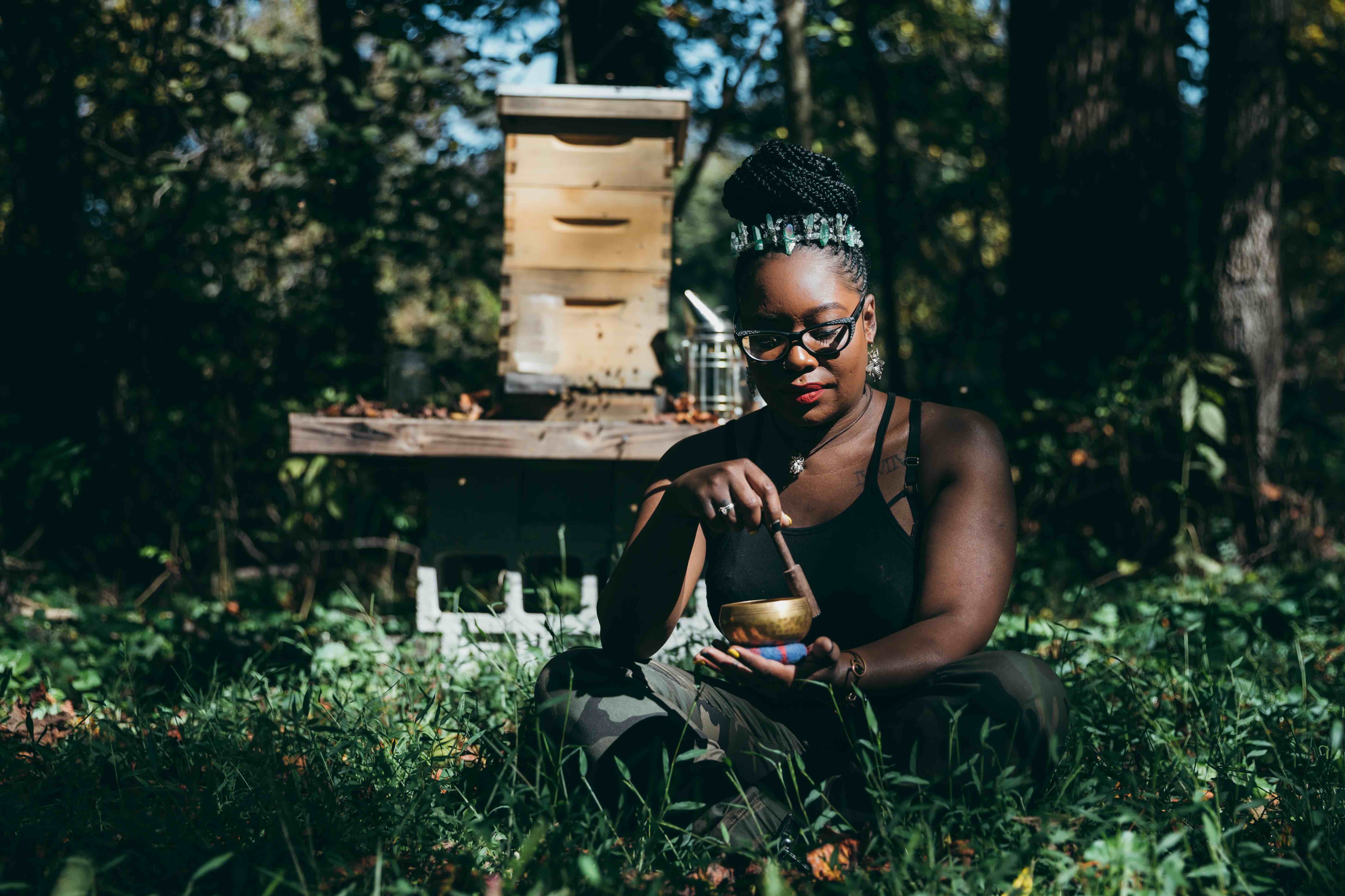 Samantha Foxx crouches in a garden holding a bowl near beehives, demonstrating her work in sustainable agriculture and beekeeping.