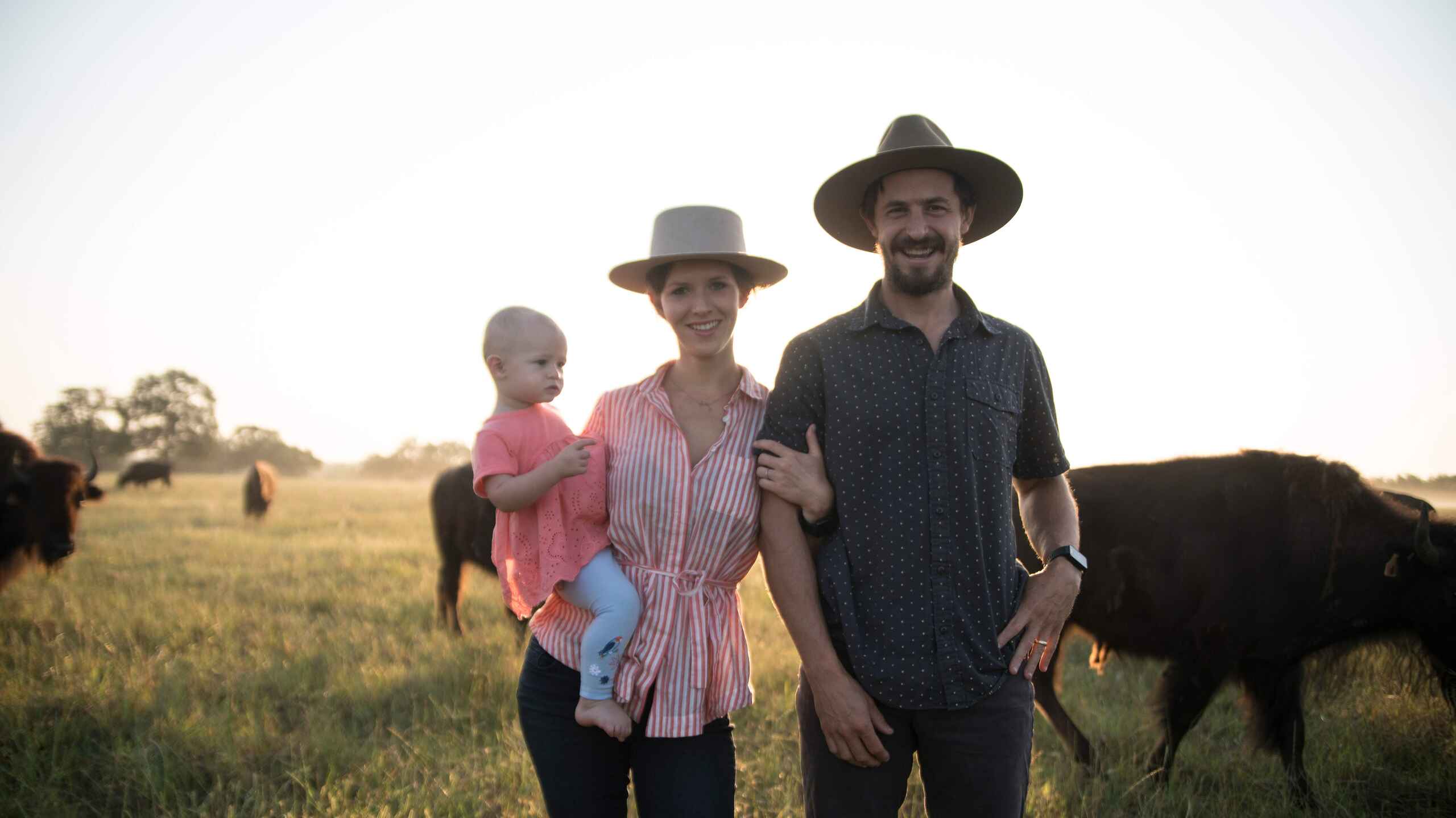 Taylor and Katie Collins with their young child stand in a pasture with cattle, wearing wide-brimmed hats at sunset