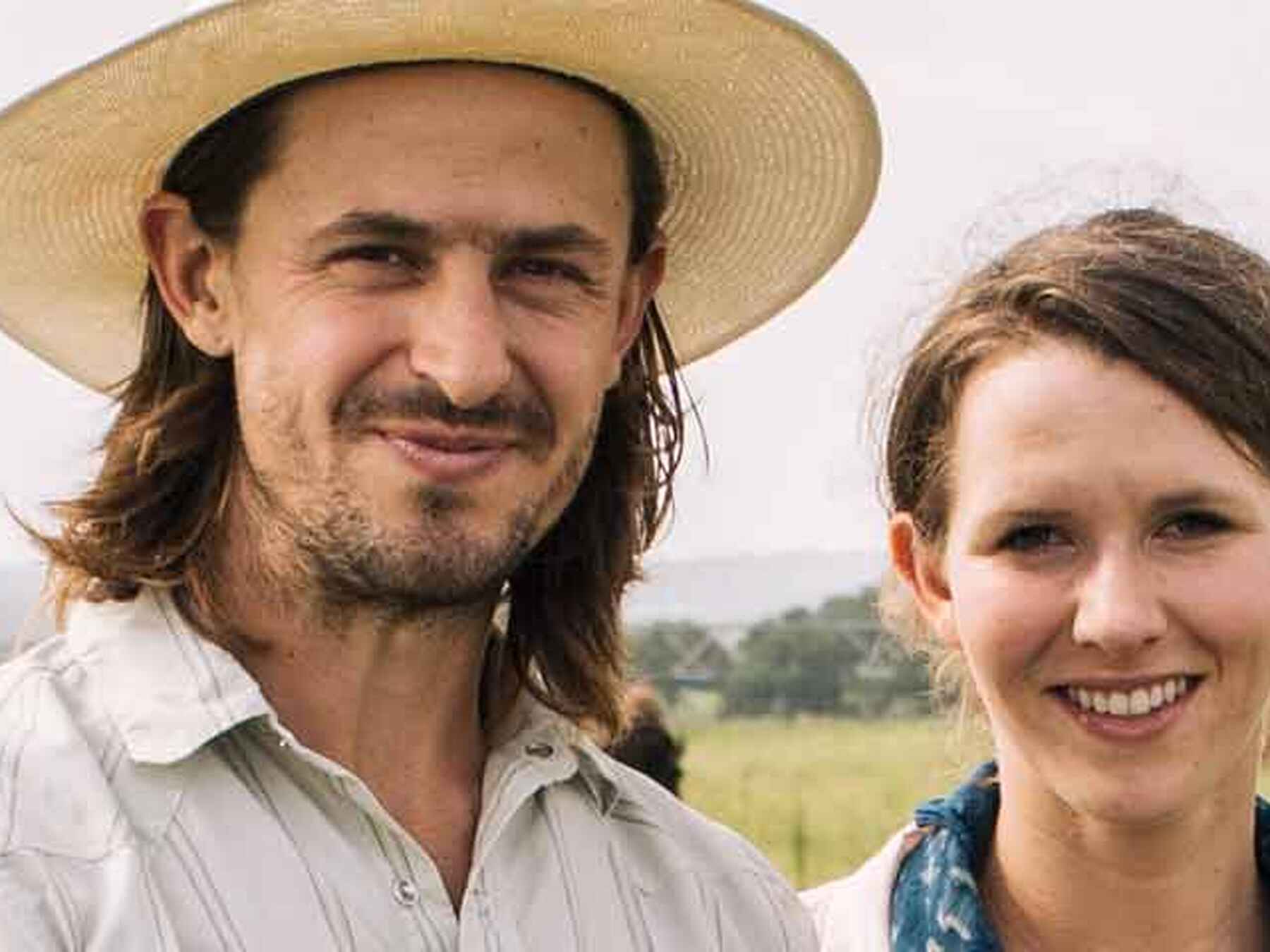 Tayloe and Katie Collins stand together in a pasture with cattle grazing in the background